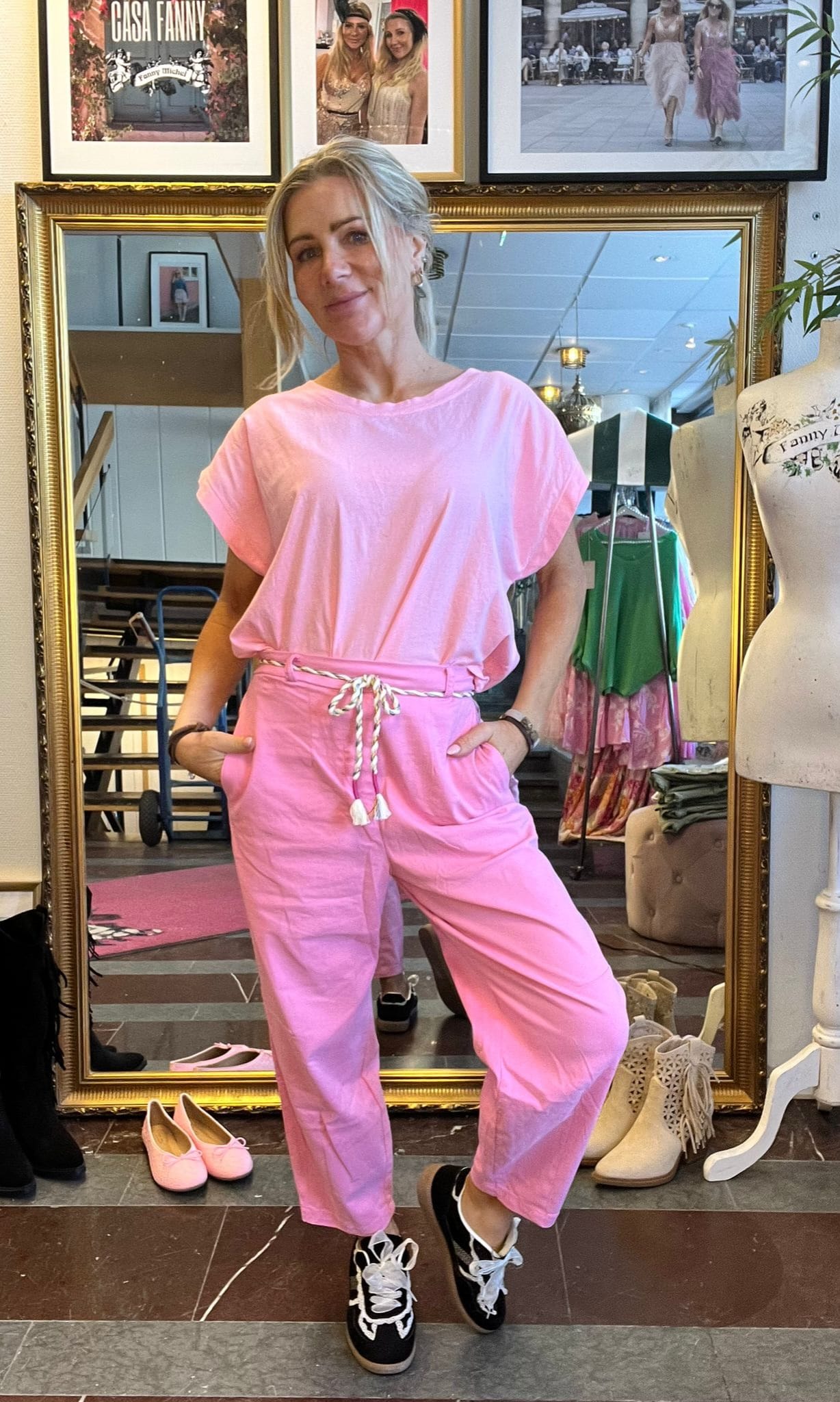 Kvinna poserar med händerna i fickorna framför en stor guldramsspegel, klädd i ljusrosa set med knytband och svarta sneakers. I klädbutiken syns skor på golvet, skyltdocka och inramade bilder med texten “CASA FANNY”.