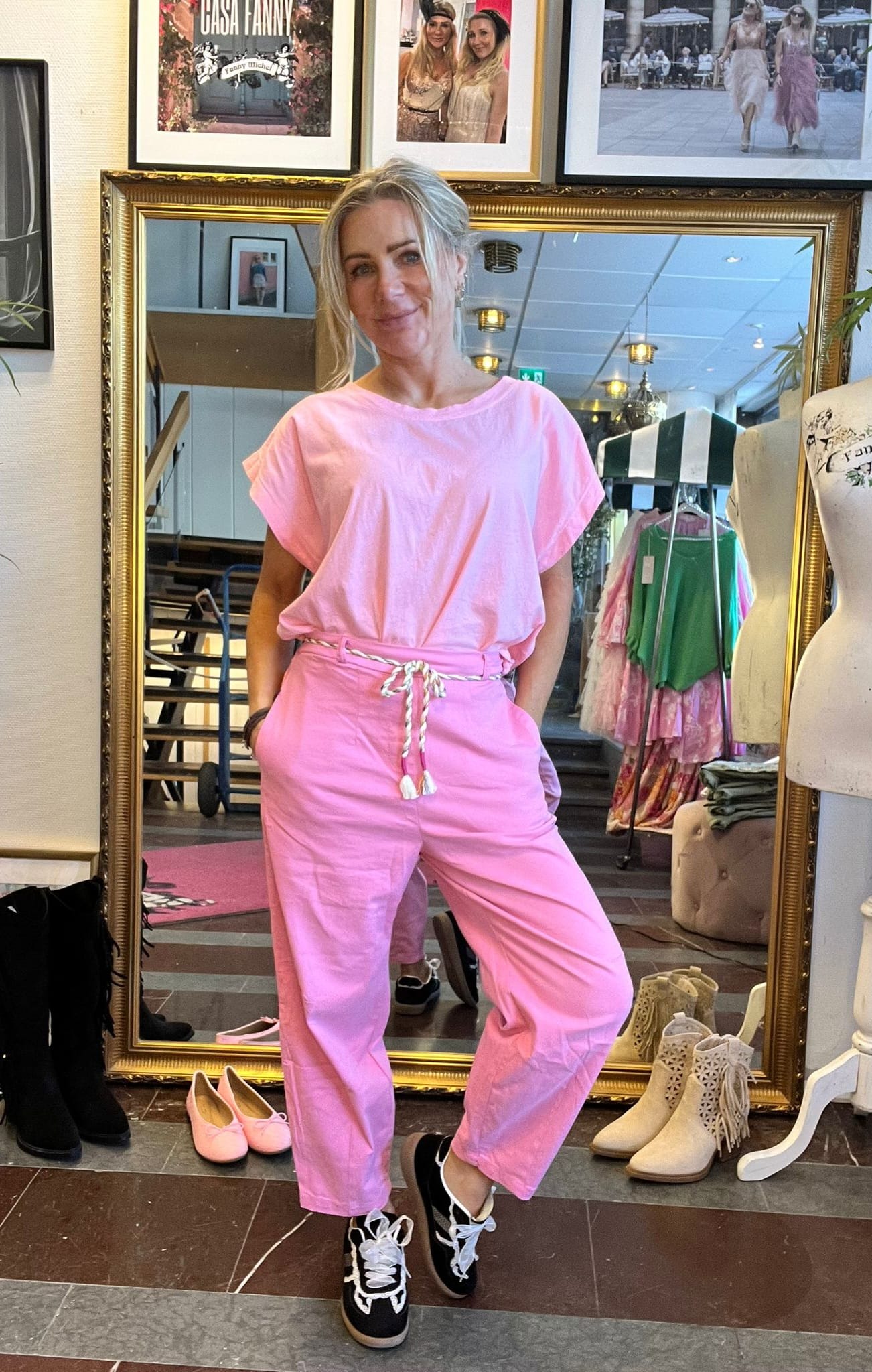 Kvinna i rosa outfit poserar framför en stor guldramsspegel i en klädbutik, med handen i fickan och ena benet fram. Runt henne syns skor, provdocka och klädhängare; på väggen står texten “CASA FANNY”.