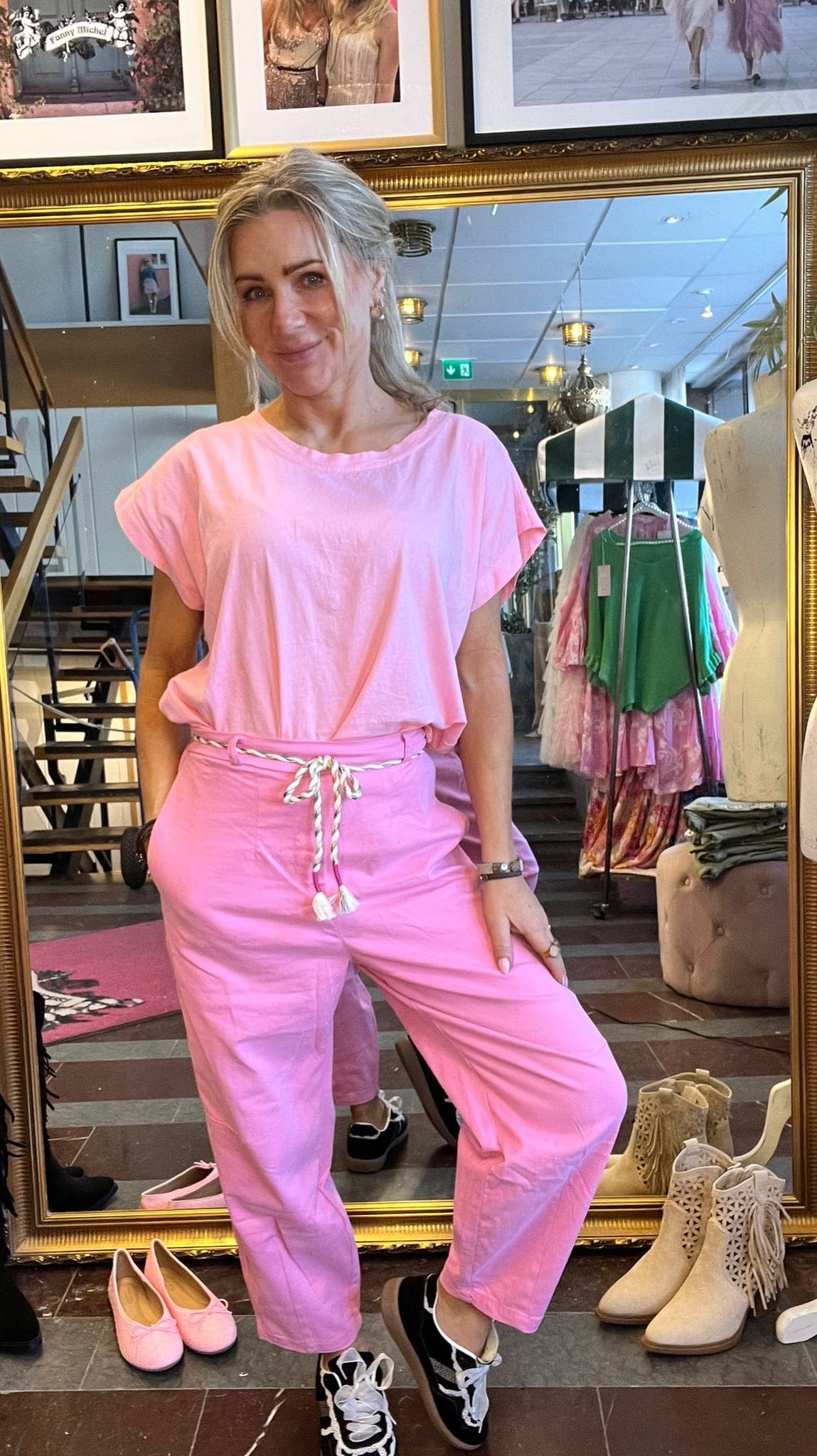 Blond kvinna poserar framför en stor guldramsspegel i en klädbutik, klädd i ljusrosa topp och rosa byxor med snörning. Hon ler med ena handen i fickan; skor och kläder syns runtom.
