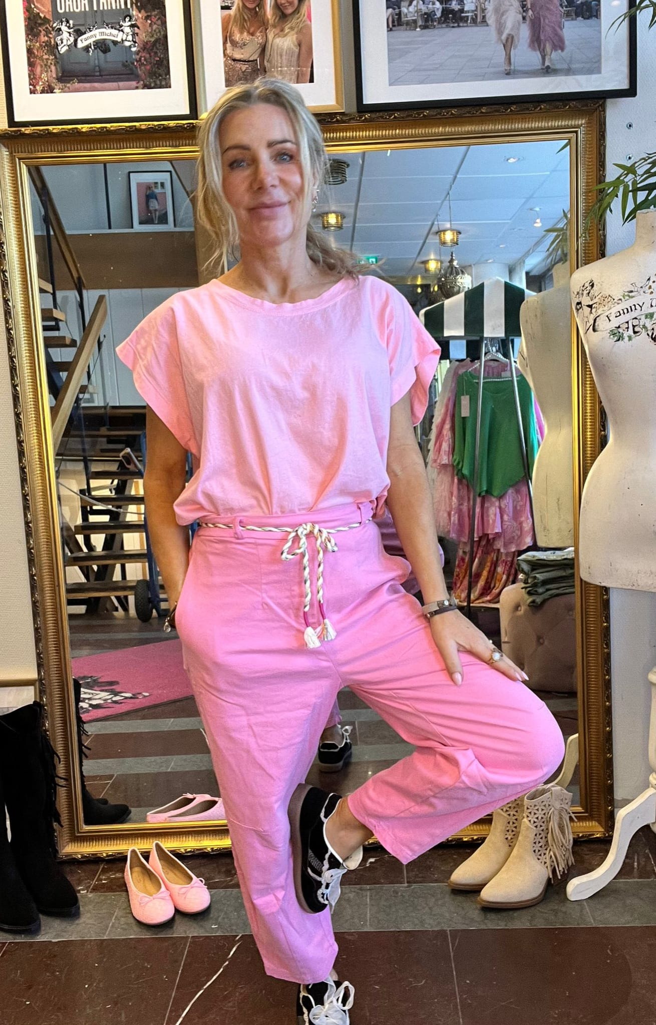 Blond kvinna poserar framför en stor guldramad spegel, klädd i rosa t-shirt och rosa byxor med snörskärp. Hon lutar ena benet på en pall; i butiken syns klädställning, skyltdocka och skor. Text: “Fanny Michel”.