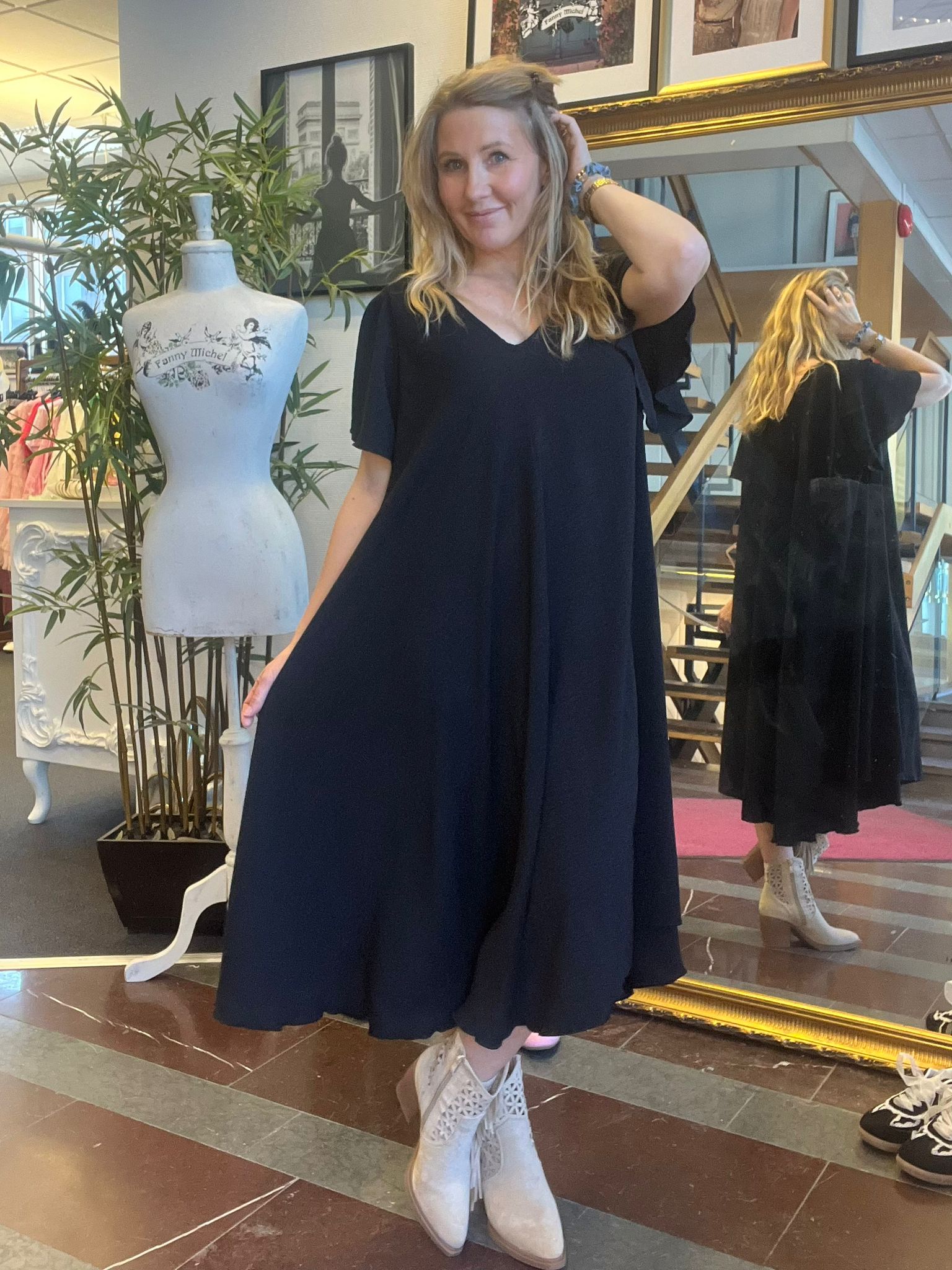Kvinna i svart, vid klänning poserar och håller upp kjolen med ena handen medan hon rör vid håret med den andra. Hon står bredvid provdocka med texten ”Fanny Michel”, spegel och trappa i butiksmiljö.