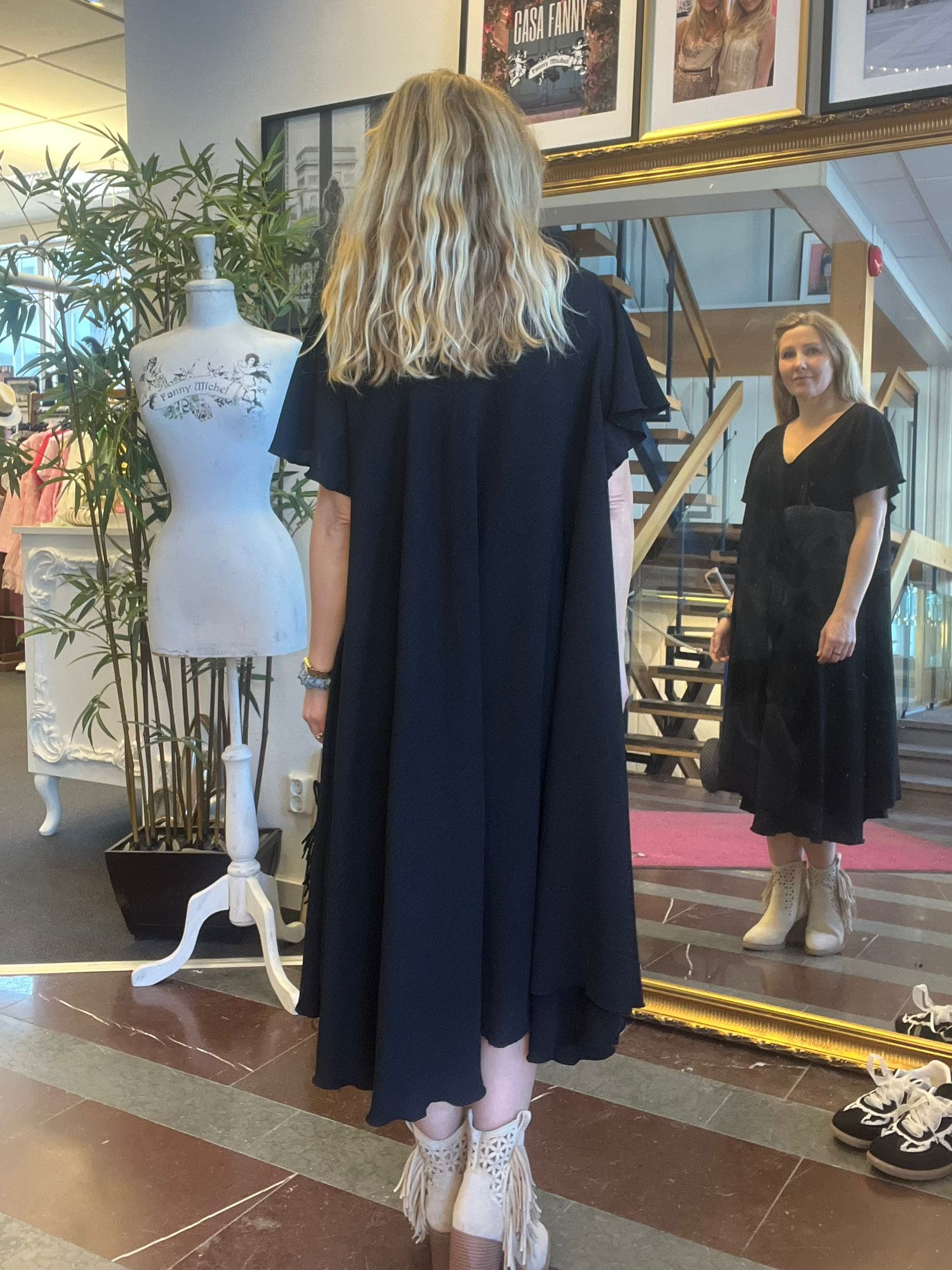 Kvinna i svart klänning och vita fransstövlar står framför en stor spegel och tittar på sin spegelbild. Bredvid finns en skyltdocka med texten “Fanny Michel” i en butiksmiljö med trappa och inramade foton.