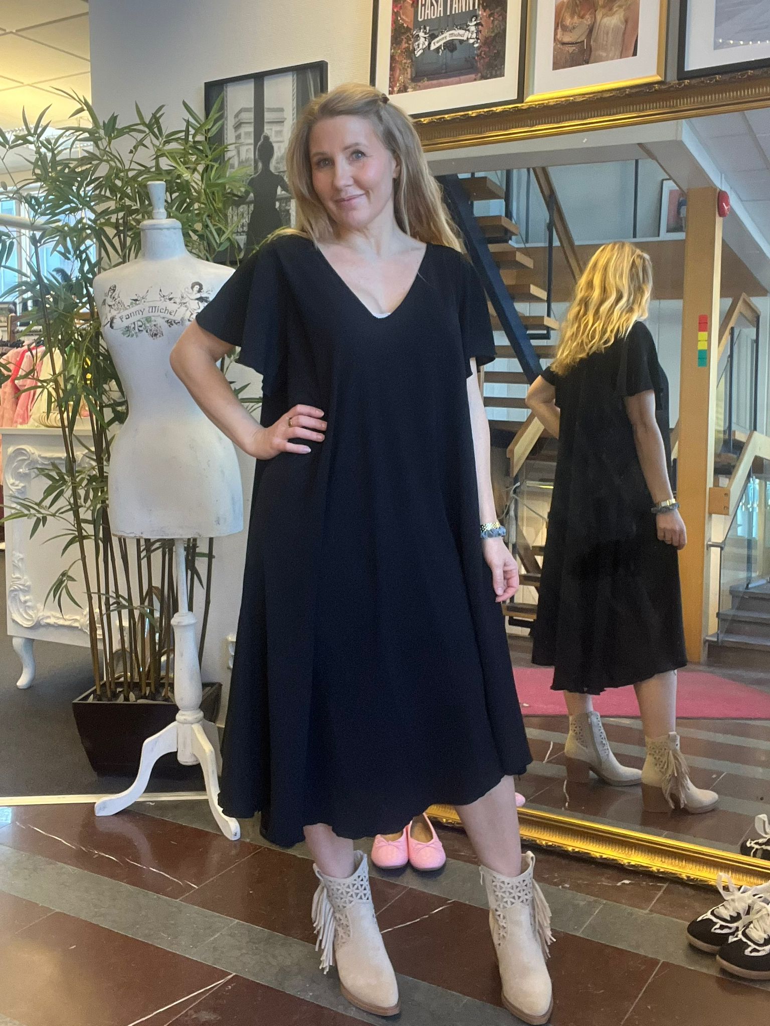 Kvinna i mörk klänning poserar med handen på höften och ler i en klädbutik eller ateljé. Bakom syns en spegel med hennes rygg, en provdocka med texten ”Fanny Michel” och en trappa.