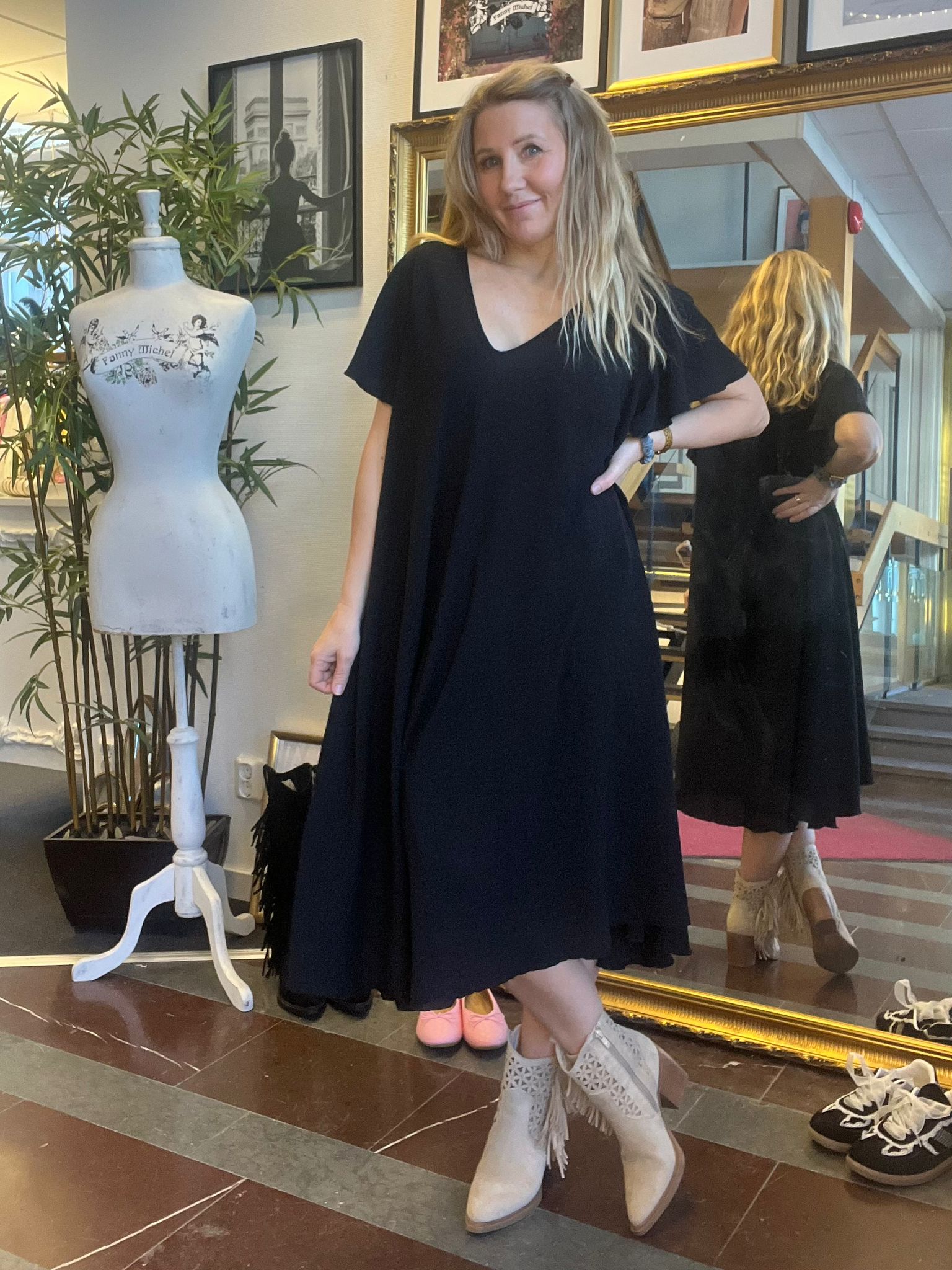 Kvinna i svart, löst sittande klänning poserar med handen på höften och ler, framför en stor guldramsspegel som visar henne bakifrån. Bredvid står en provdocka med texten “Fanny Michel” i en butiksmiljö.