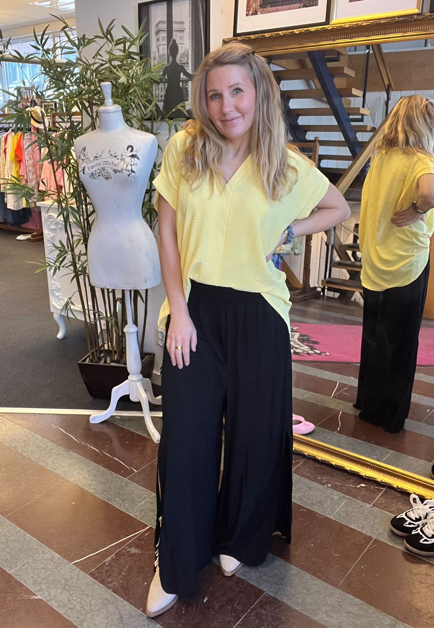 Kvinna står och poserar med handen på höften i en klädbutik, klädd i gul topp och svarta byxor. Bredvid finns en provdocka med texten ”Funny Mischel” och en stor spegel som visar hennes rygg.