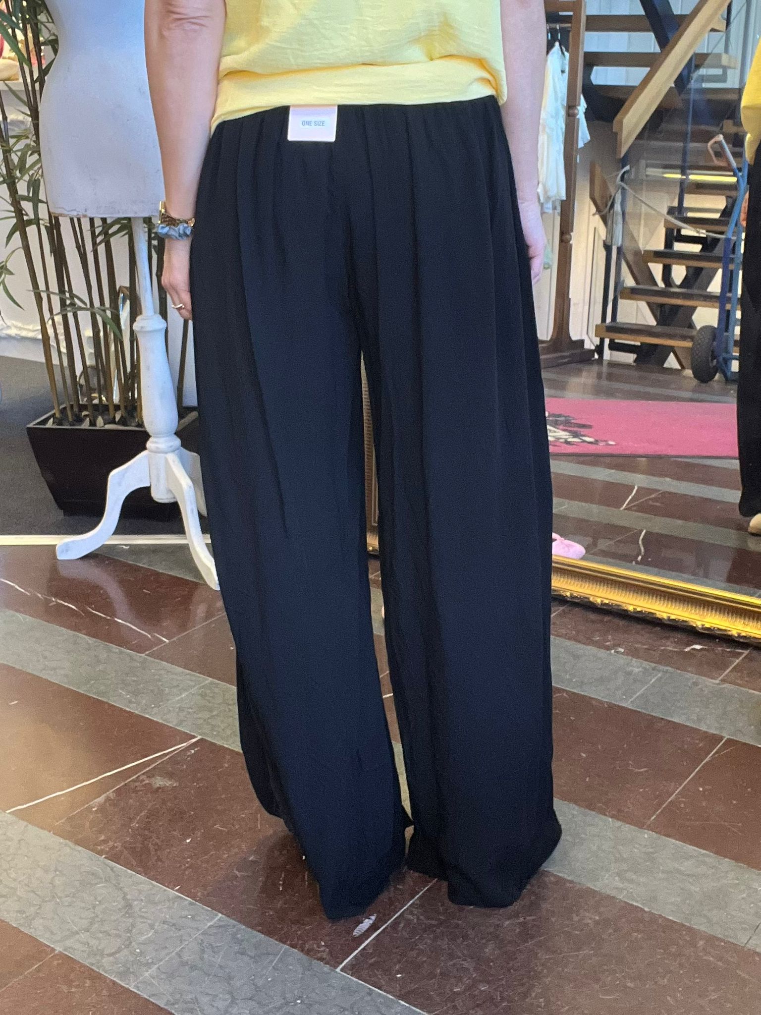Person står i en butik och provar svarta, vida byxor med hög midja och veck framtill. En etikett vid midjan visar texten “ONE SIZE”. Gul topp syns ovanför byxlinningen; spegel och trappa i bakgrunden.