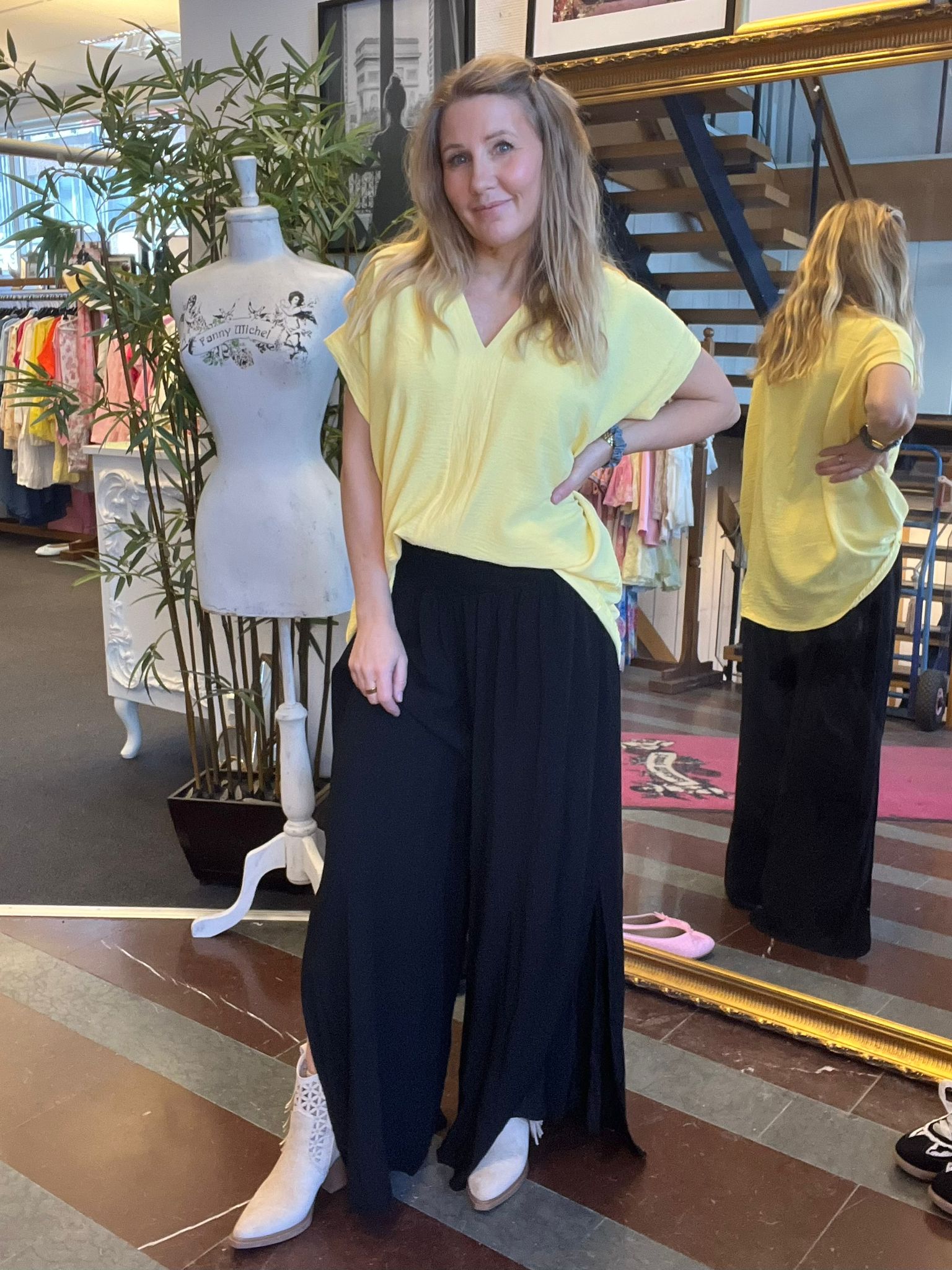 Kvinna står och poserar med handen på höften i en klädbutik, iklädd gul topp, svart vidbyxa och vita boots. En stor spegel visar ryggen; intill syns provdocka med texten “Fanny Michel” och klädställningar.
