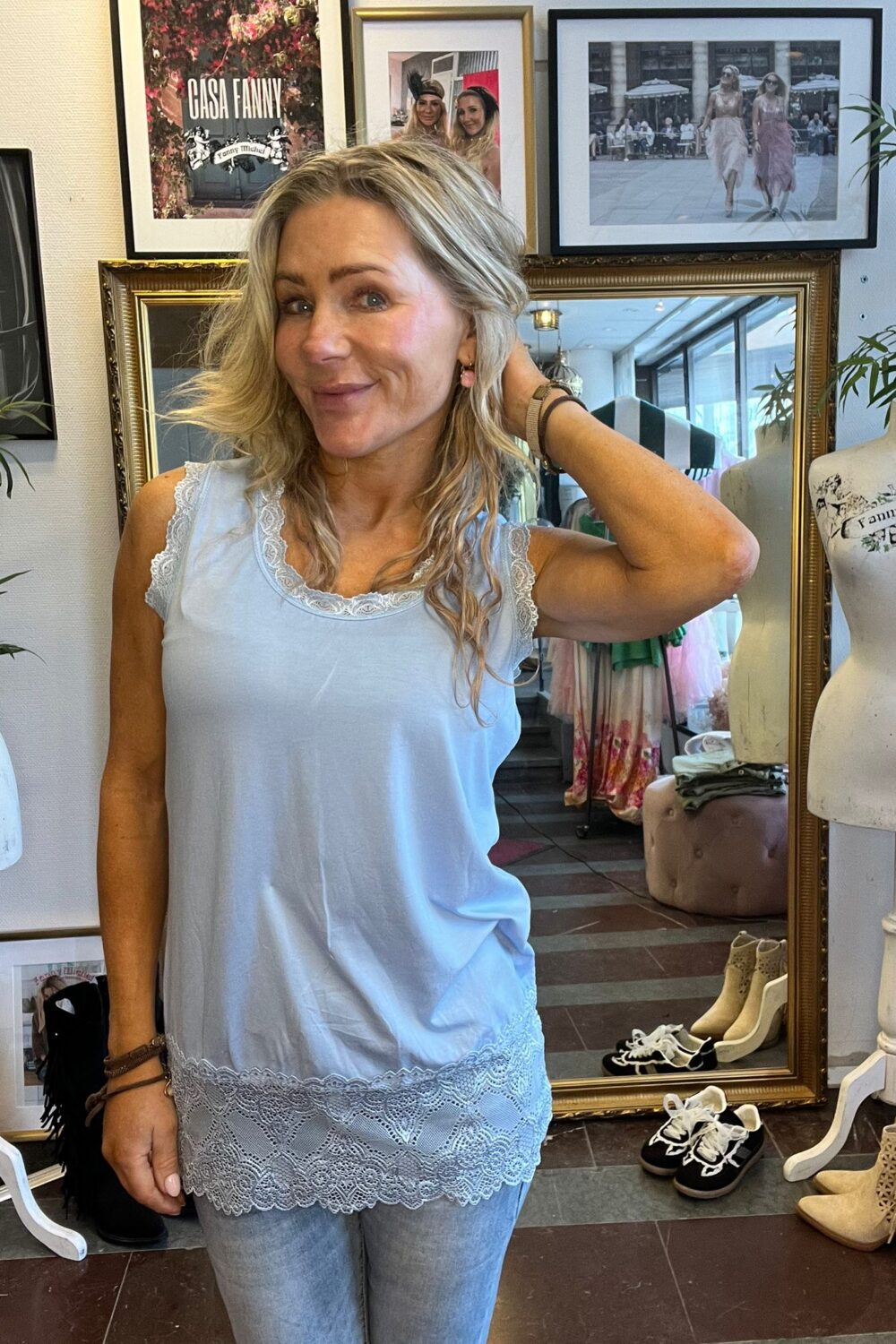 Leende kvinna i ljusblå spetslinne poserar med ena handen i håret i en klädbutik. Bakom henne syns en stor spegel, skyltdockor, skor på golvet och inramade foton med texten ”CASA FANNY”.