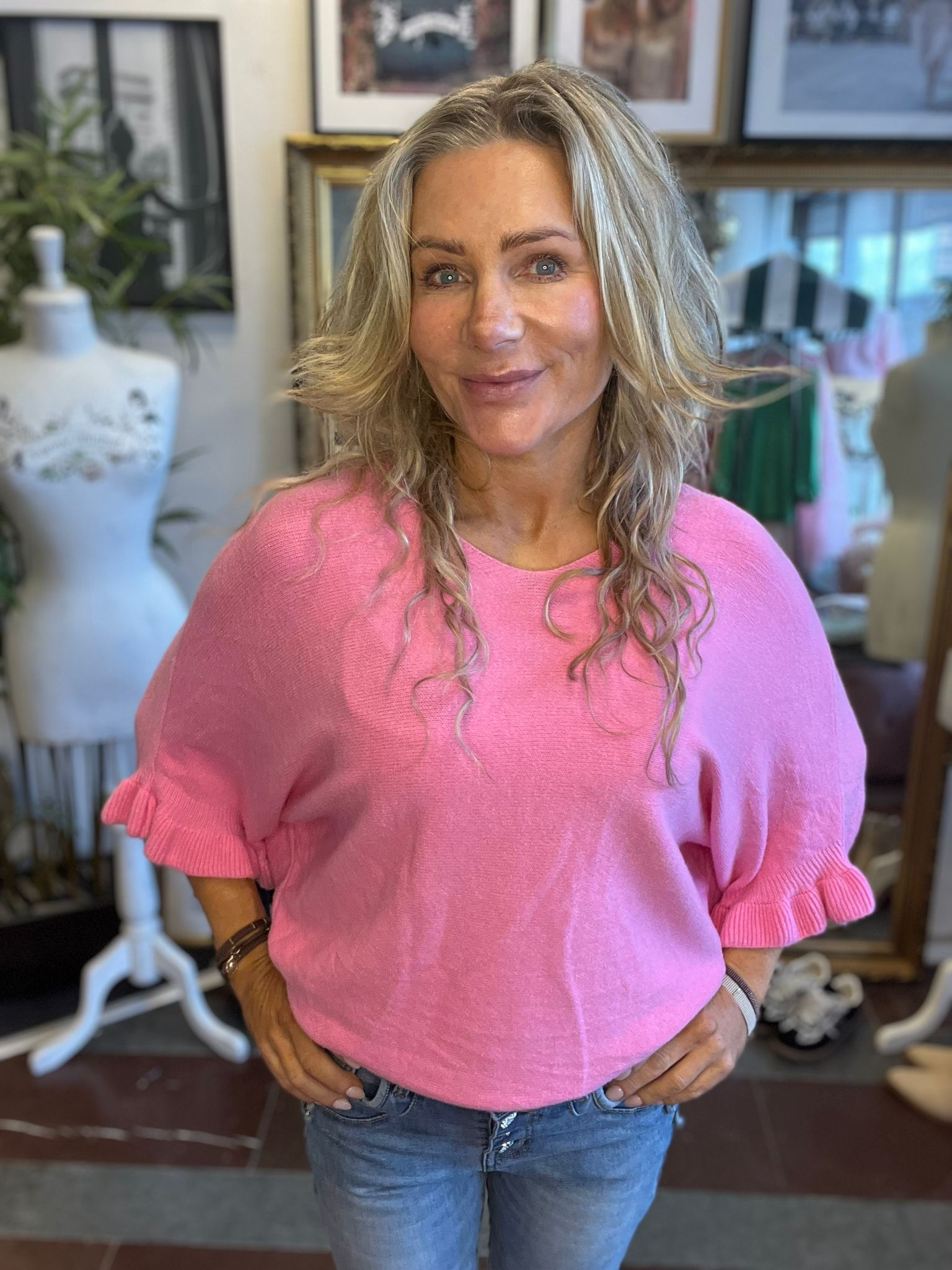 Leende kvinna med långt blont hår står med händerna i jeansfickorna och tittar in i kameran. Hon bär en rosa stickad tröja. I bakgrunden syns klädskyltdockor, speglar och inramade bilder i en butik.