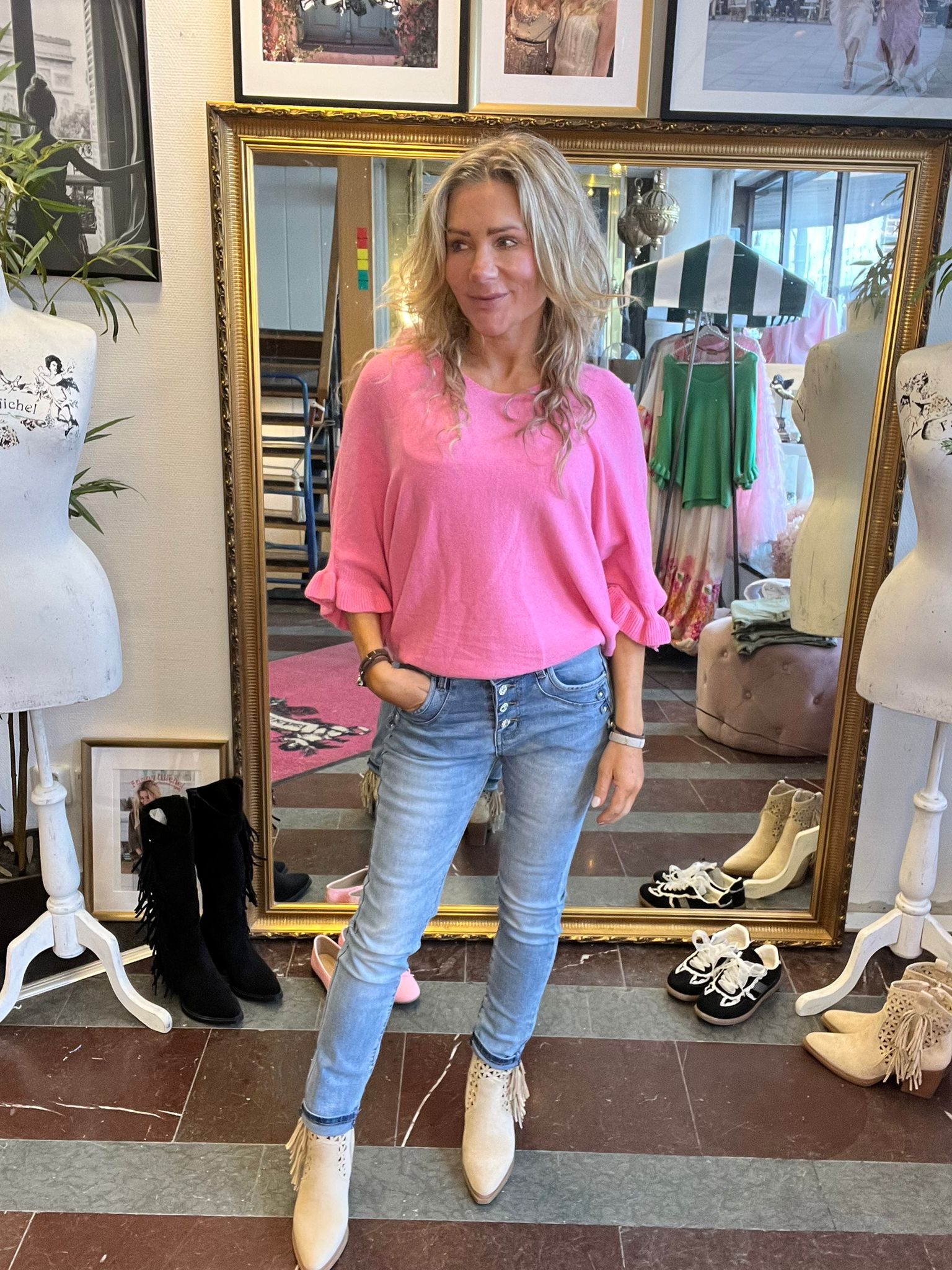 Blond kvinna i rosa tröja och ljusa jeans står med ena handen i fickan och tittar åt sidan framför en stor guldramsspegel i en klädbutik, omgiven av provdockor, klädställning och skor på golvet.