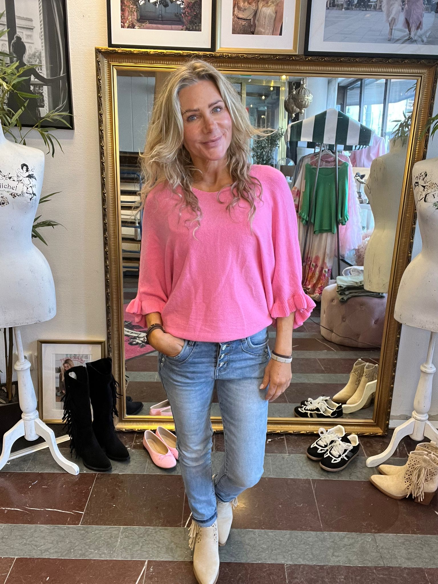 Kvinna i rosa tröja och ljusa jeans står avslappnat med en hand i fickan och ler mot kameran, framför en stor guldramsspegel i en klädbutik med klädställning, skyltdockor och skor på golvet.