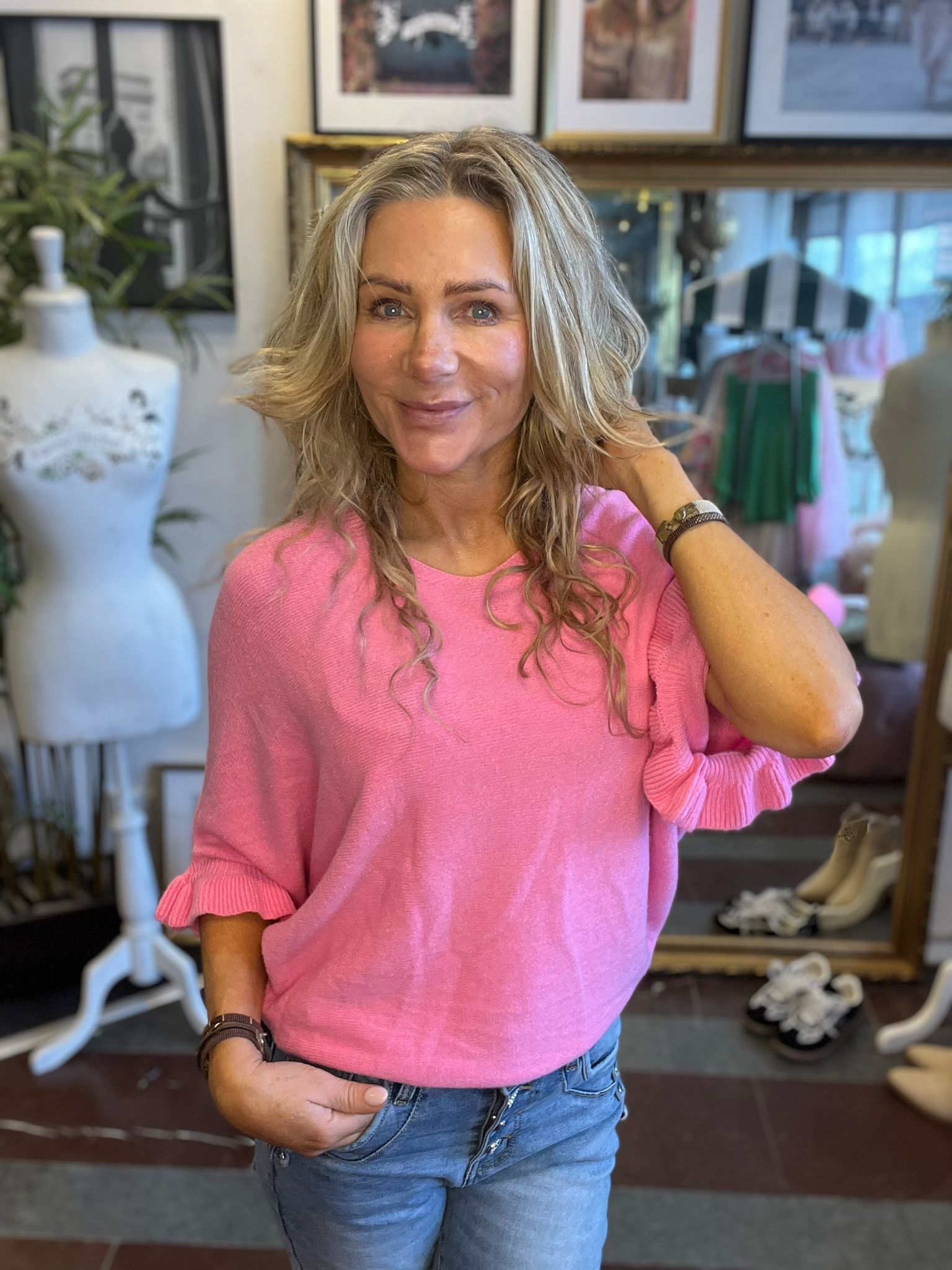 Kvinna med långt blont hår ler mot kameran och håller en hand bakom nacken. Hon bär rosa tröja och jeans. I bakgrunden syns en butiksmiljö med provdocka, kläder på ställning och inramade bilder.