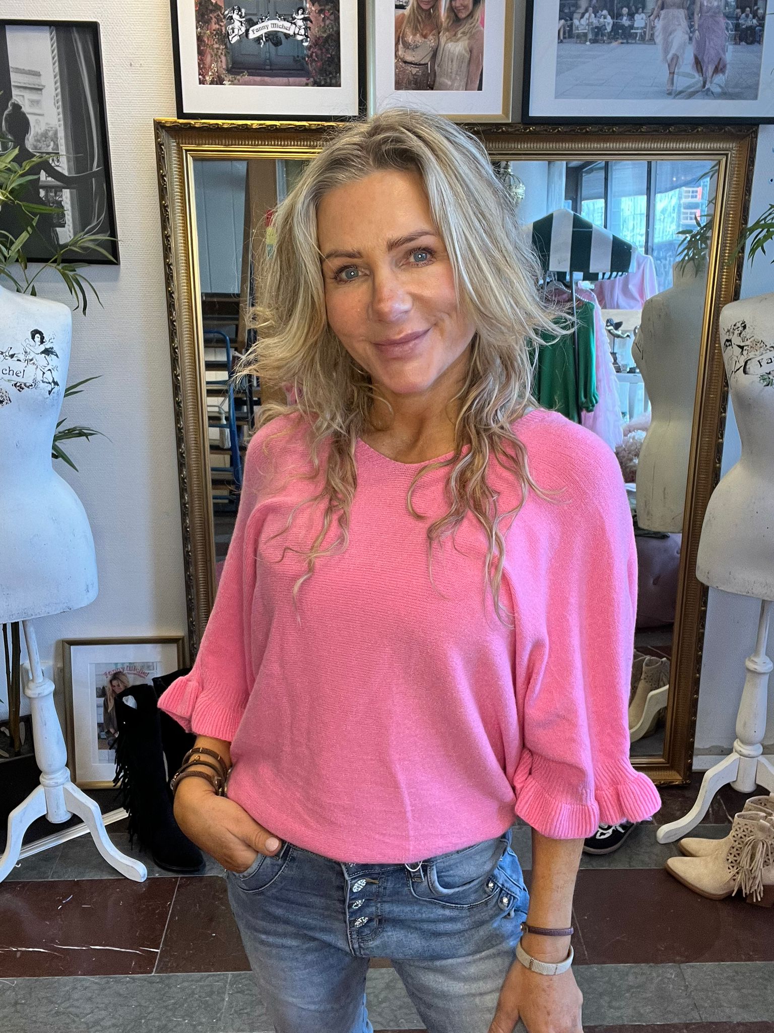 Leende kvinna med långt blont hår står med handen i jeansfickan och tittar mot kameran, klädd i rosa tröja. Bakom henne syns en stor guldramsspegel, provdockor och inramade foton i en butik/ateljémiljö.