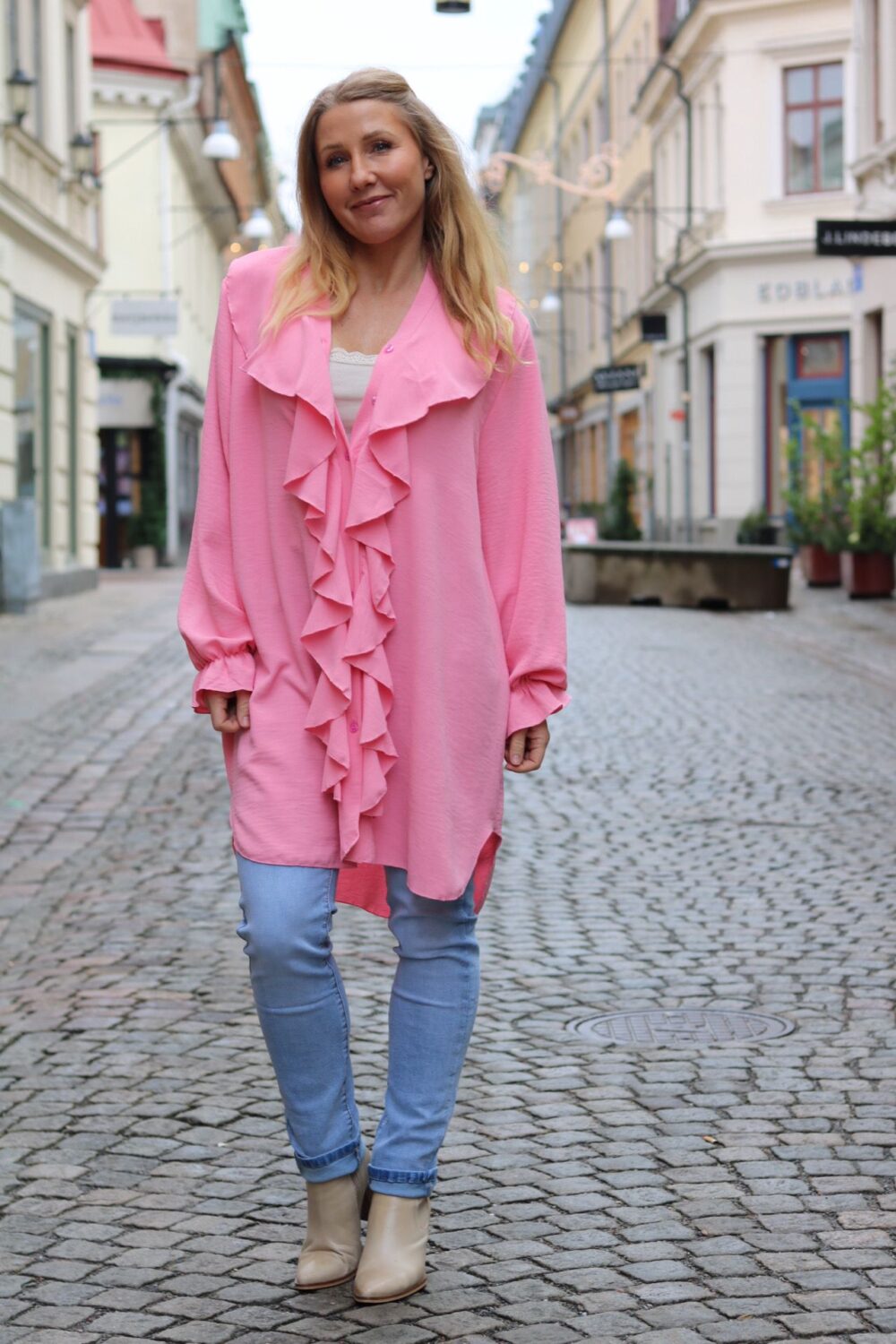 Kvinna med långt blont hår står och ler på en kullerstensgata, klädd i en ljusrosa volangtunika över vit topp, ljusa jeans och beige boots. Bakom syns smala stadshus, gatlyktor och butiksfasader.