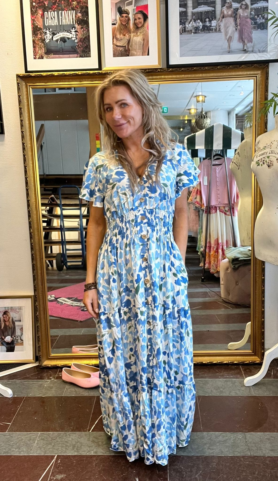 Kvinna poserar och ler framför en stor guldramsspegel, iklädd lång blåvit blommig klänning med knappar. I bakgrunden syns en butik med kläder, provdocka och inramade foton; texten “CASA FANNY”.