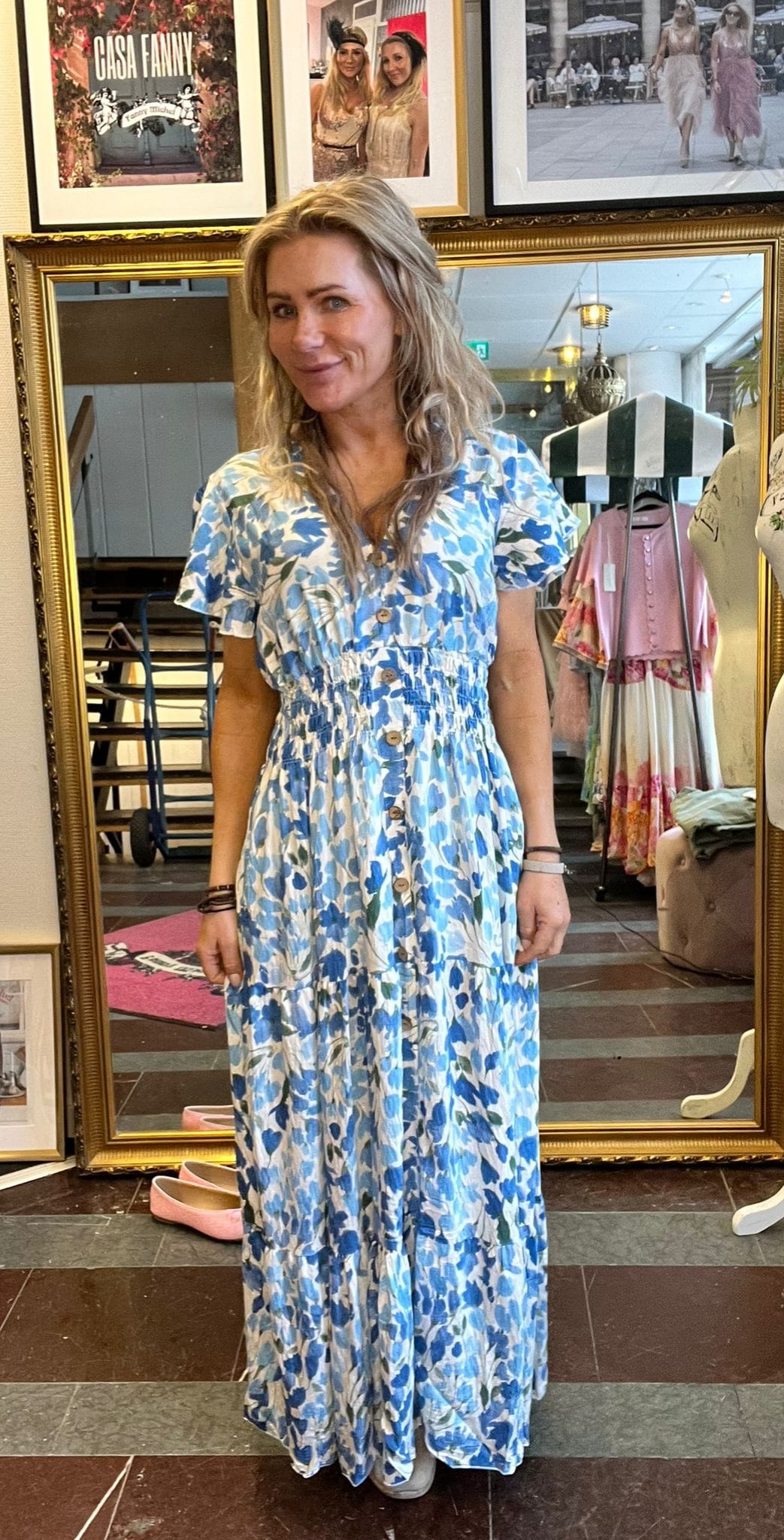 Kvinna står och ler framför en stor guldramsspegel i en klädbutik, iklädd blåvit blommig maxiklänning med knappar. I bakgrunden syns klädställning och inramade foton med texten “CASA FANNY”.