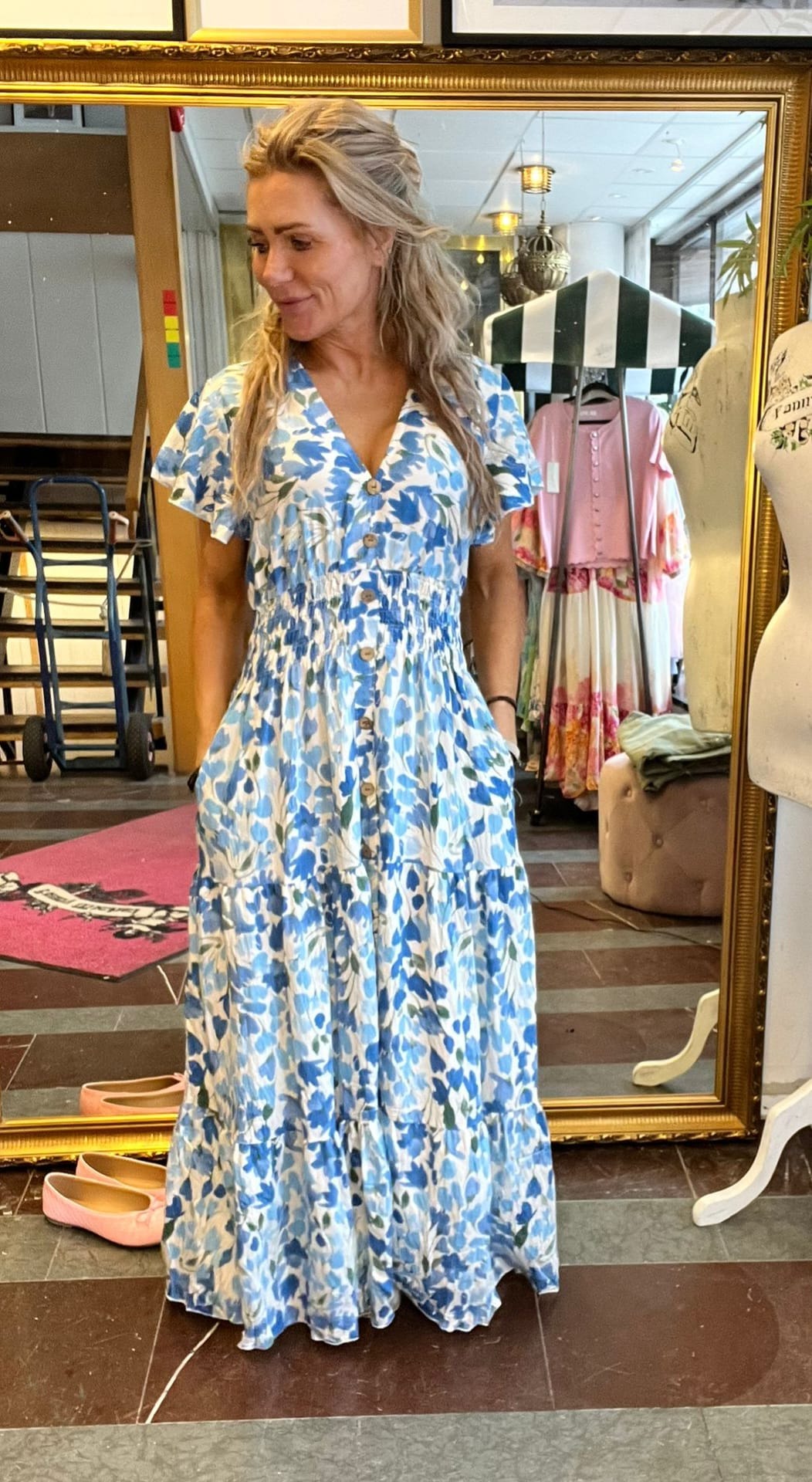 Kvinna med långt blont hår provar en blåvit blommig maxiklänning med knappar och fickor, och tittar åt sidan med ett litet leende. Hon står framför en stor guldramsspegel i en klädbutik med klädställning bakom.