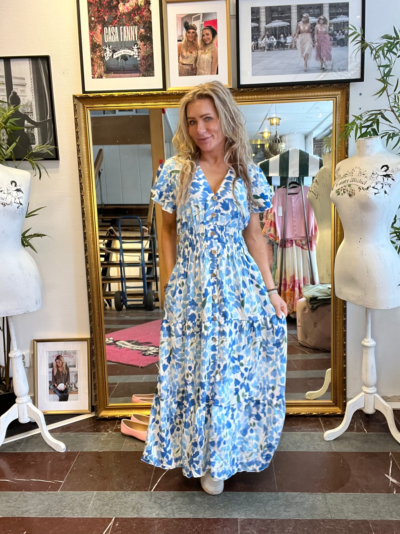 Kvinna i blåvit blommig maxiklänning står och poserar framför en stor guldramsspegel i en butik. Runt henne syns provdockor och inramade foton på väggen, bland annat texten “CASA FANNY”.