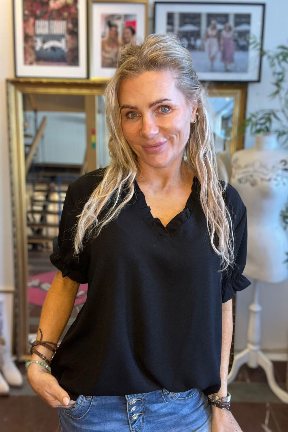 Leende kvinna med långt blont hår och svart blus står med handen i jeansfickan och tittar mot kameran. I bakgrunden syns en guldramsspegel, inramade fotografier på väggen och en provdocka i en ateljélik miljö.