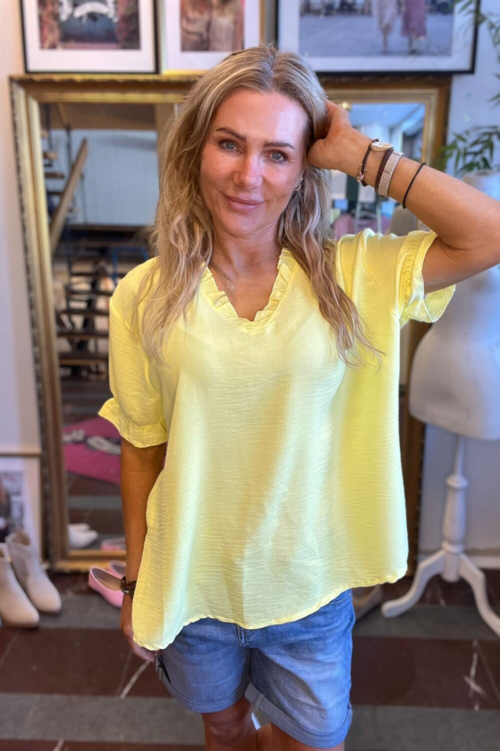 Kvinna med långt blont hår ler mot kameran och håller ena handen mot huvudet. Hon bär ljusgul volangblus och blå shorts. Står i ett rum med stor guldramad spegel, skor på golvet och en provdocka.