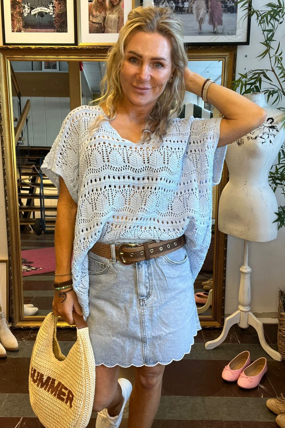 Leende blond kvinna står och poserar med ena handen bakom huvudet och håller en halmstrandväska. Hon bär ljusblå virkad topp och ljus jeanskjol, i en butik med speglar, inramade foton och skyltdocka. Text i tavla: ”Fanny Michel”.