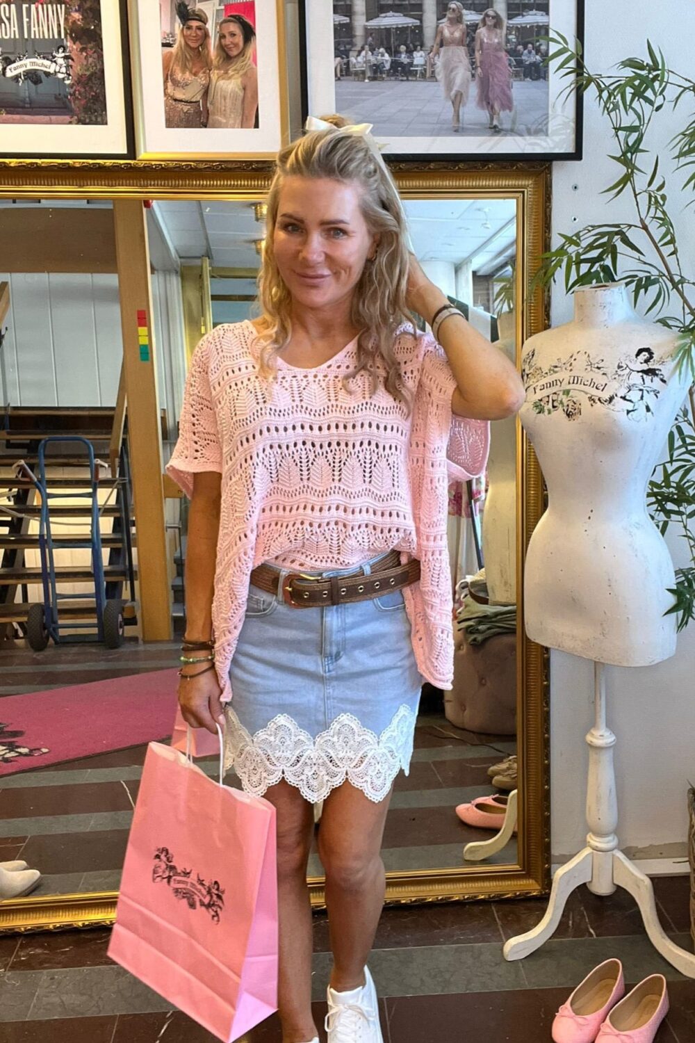 Leende kvinna står i en butik eller studio och håller en rosa shoppingpåse, med ena handen i håret. Hon bär rosa virkad topp och ljus kjol med spetskant; bakom syns spegel, inramade foton och provdocka med texten ”Fanny Michel”.