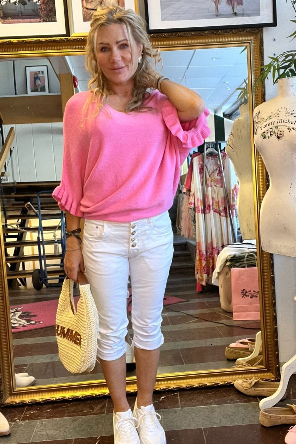 Blond kvinna poserar framför en stor guldramsspegel i en klädbutik, med ena handen i håret och en stickad väska i den andra. Hon bär rosa tröja, vita capribyxor och vita sneakers; skylten “CASA FANNY” syns.