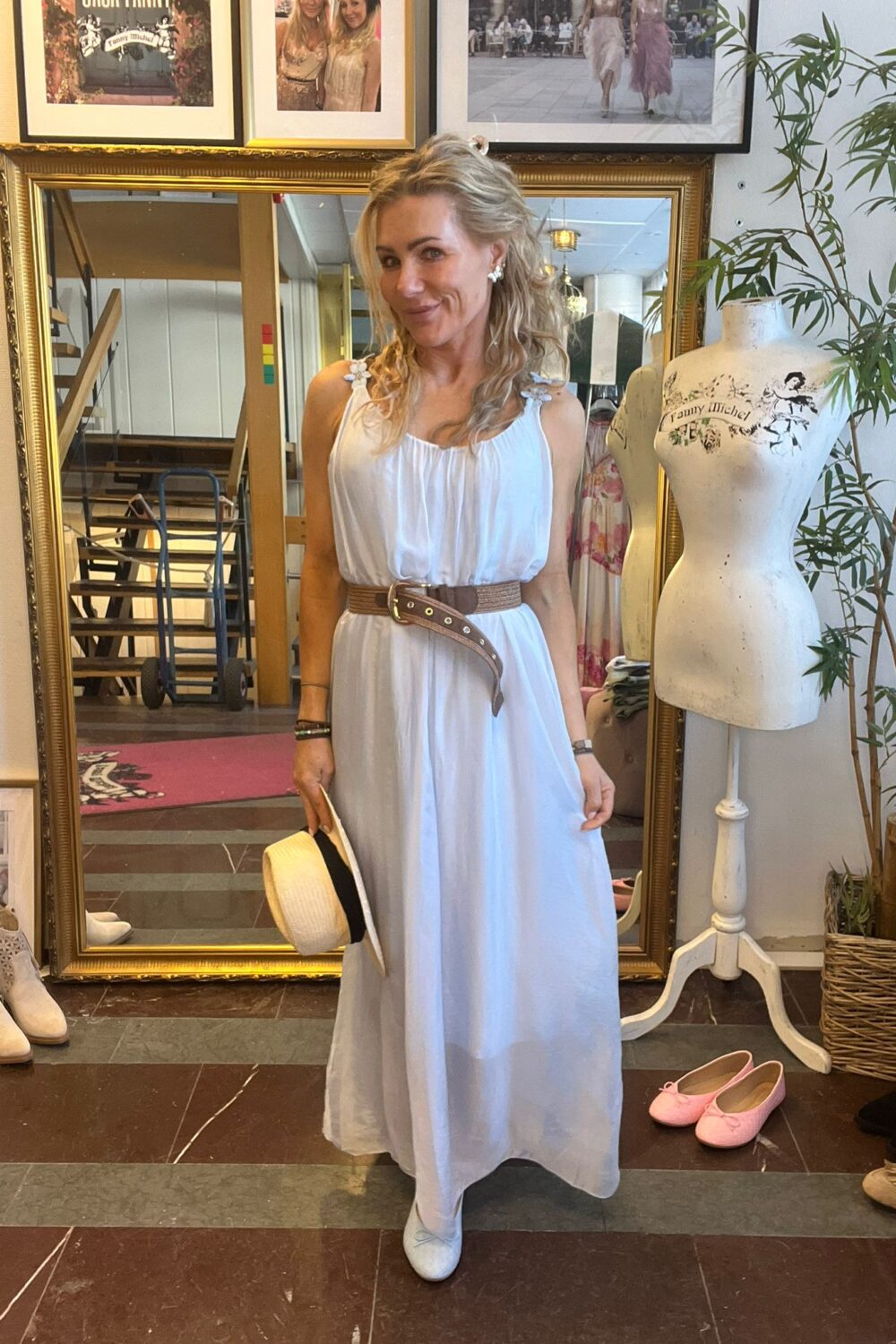 Leende kvinna i vit, luftig klänning med brunt bälte håller en stråhatt och poserar framför en stor guldramsspegel i en butik. Bredvid står en provdocka med texten “Fanny Michel”, och skor ligger på golvet.