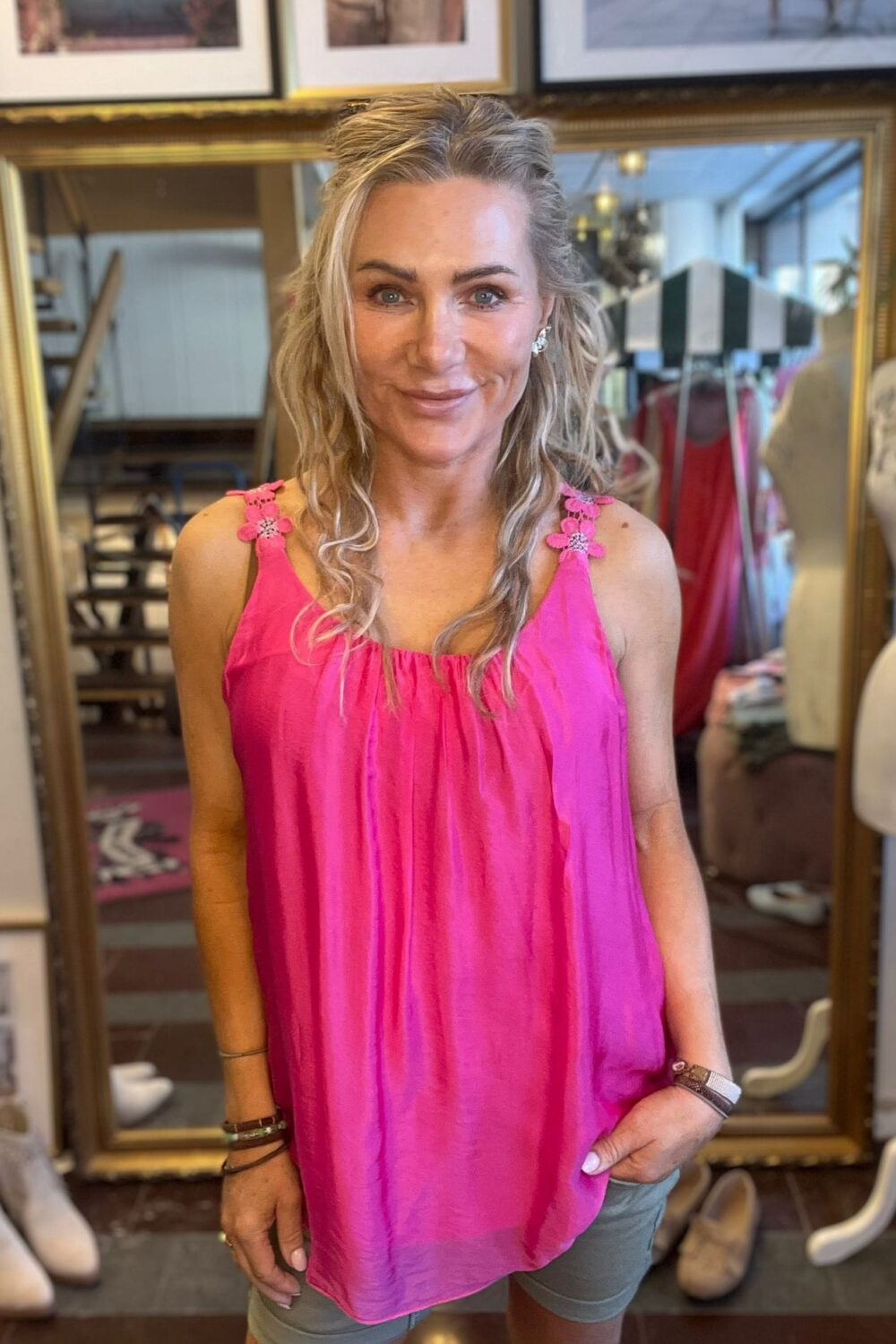 Leende kvinna med vågigt blont hår står och poserar framför en stor guldramsspegel, klädd i ceriserosa linne med blommiga axelband och grå shorts. I bakgrunden syns en butik med klädställningar och inramade foton.