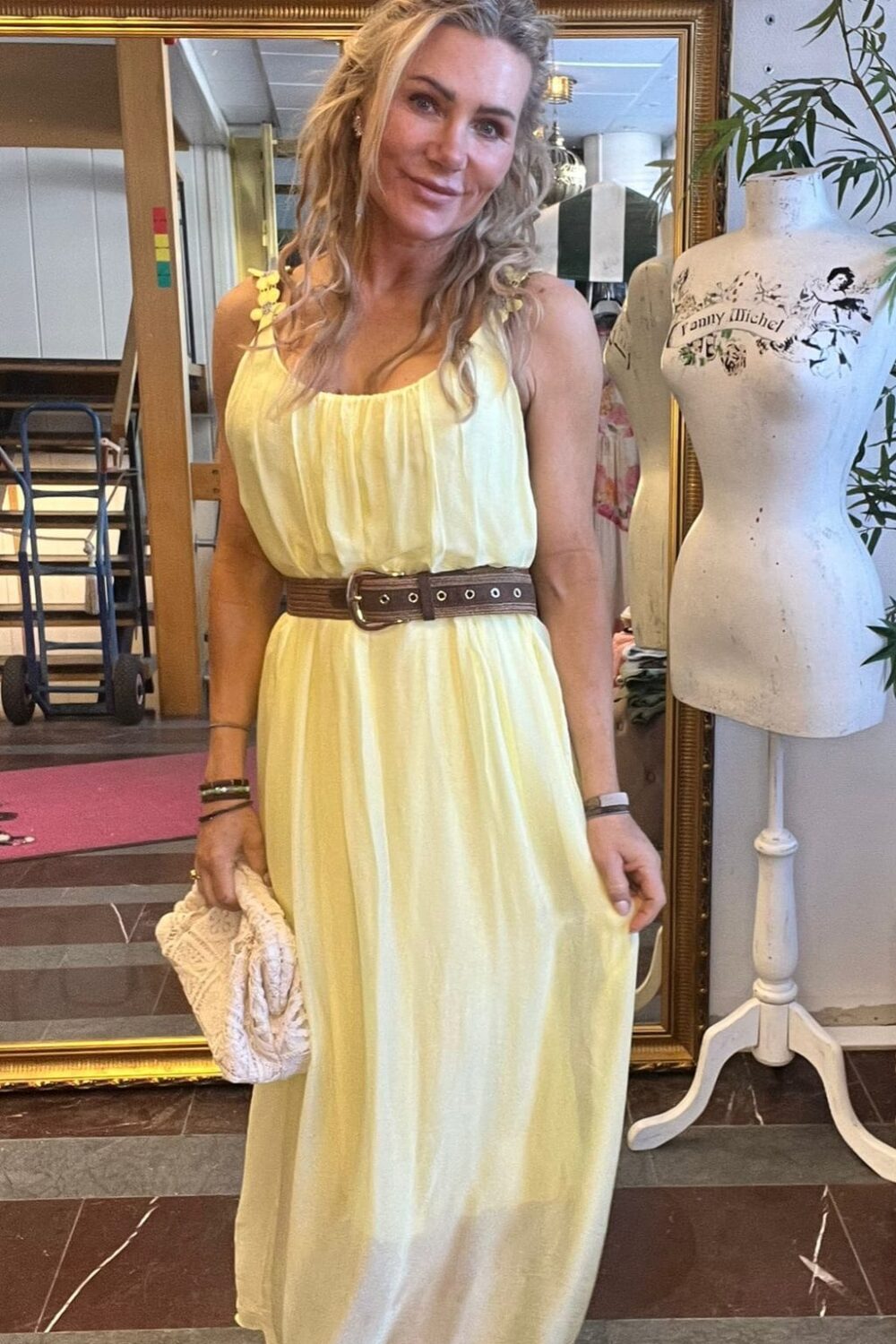 Leende kvinna i lång ljusgul klänning med brunt bälte står inomhus och håller en beige virkad väska. Bakom henne syns spegel, inramade foton och en provdocka med texten “Fanny Michel”.