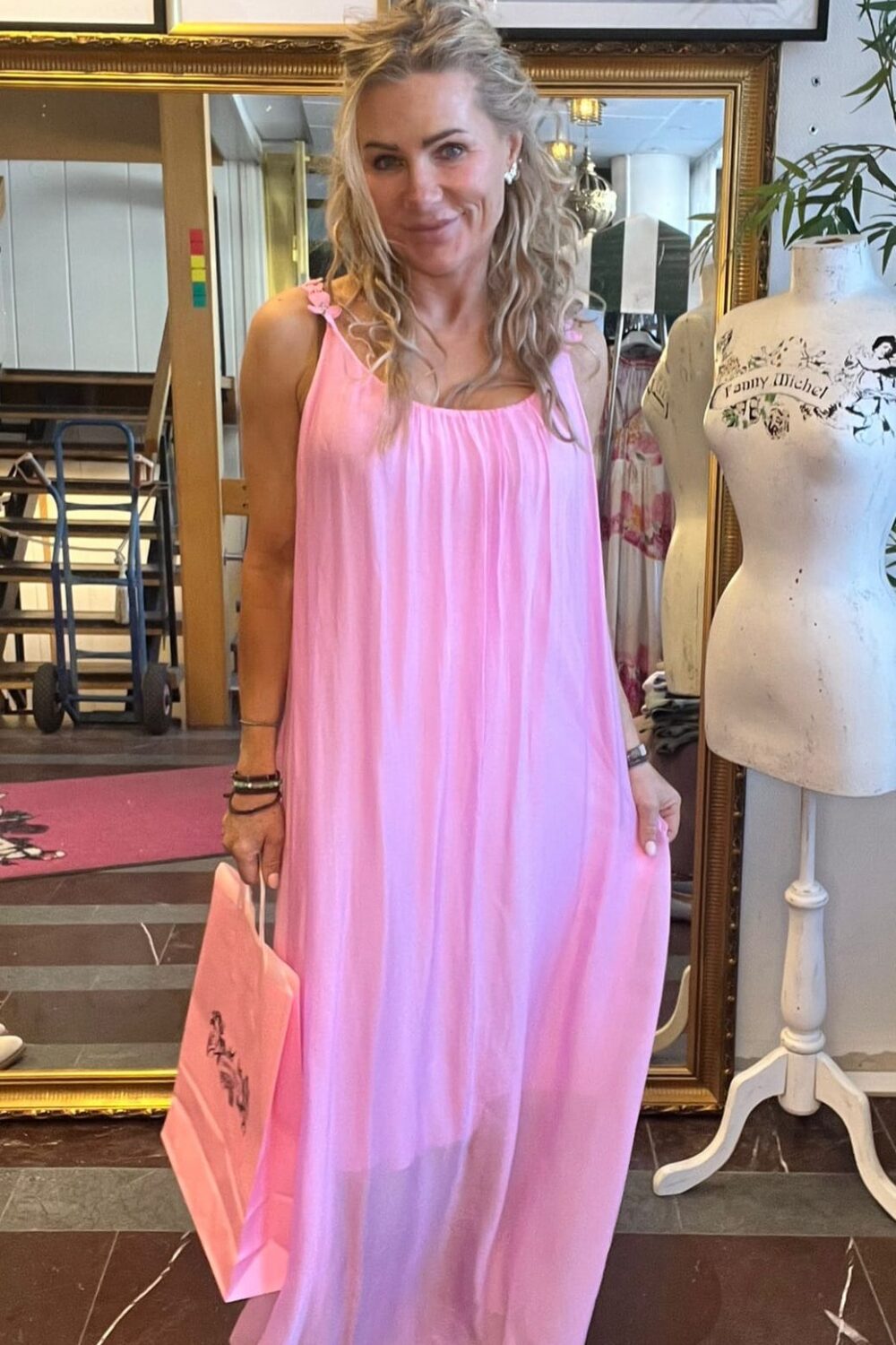 Leende kvinna i lång, rosa klänning står i en butik och håller en rosa shoppingpåse. Bredvid syns en provdocka med texten “Fanny Michel”, samt inramade modefoton och en stor spegel i bakgrunden.
