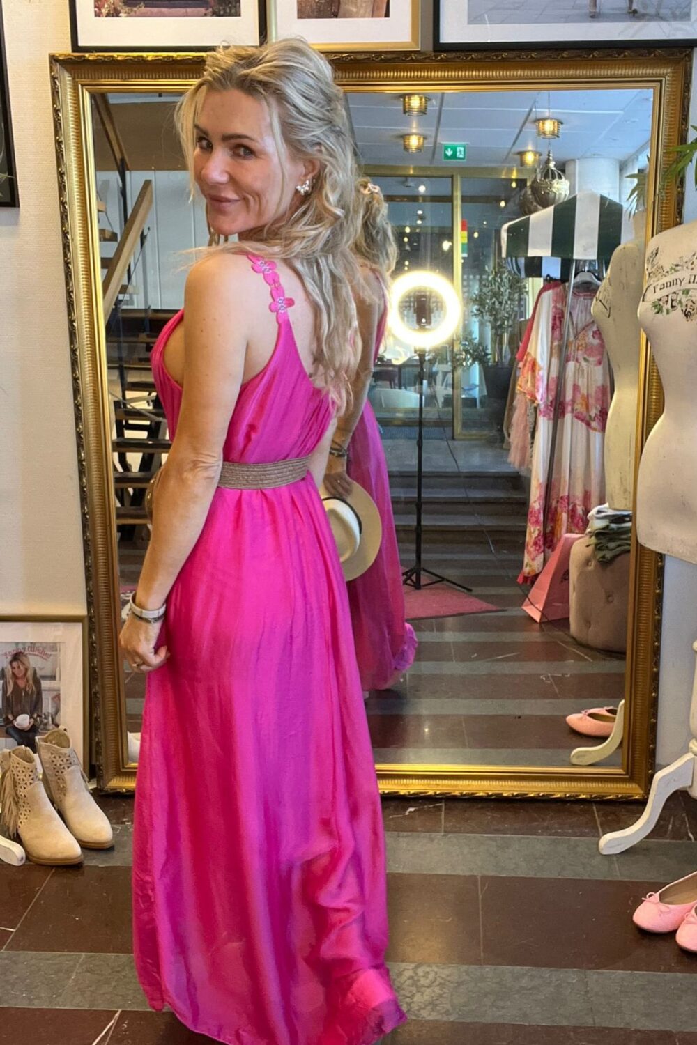 Kvinna i fuchsiarosa långklänning vänder sig om och ler mot kameran framför en stor guldramsspegel i en butik. I spegeln syns ringljus, klädställning och inredning; runtom står skor och skyltdocka.