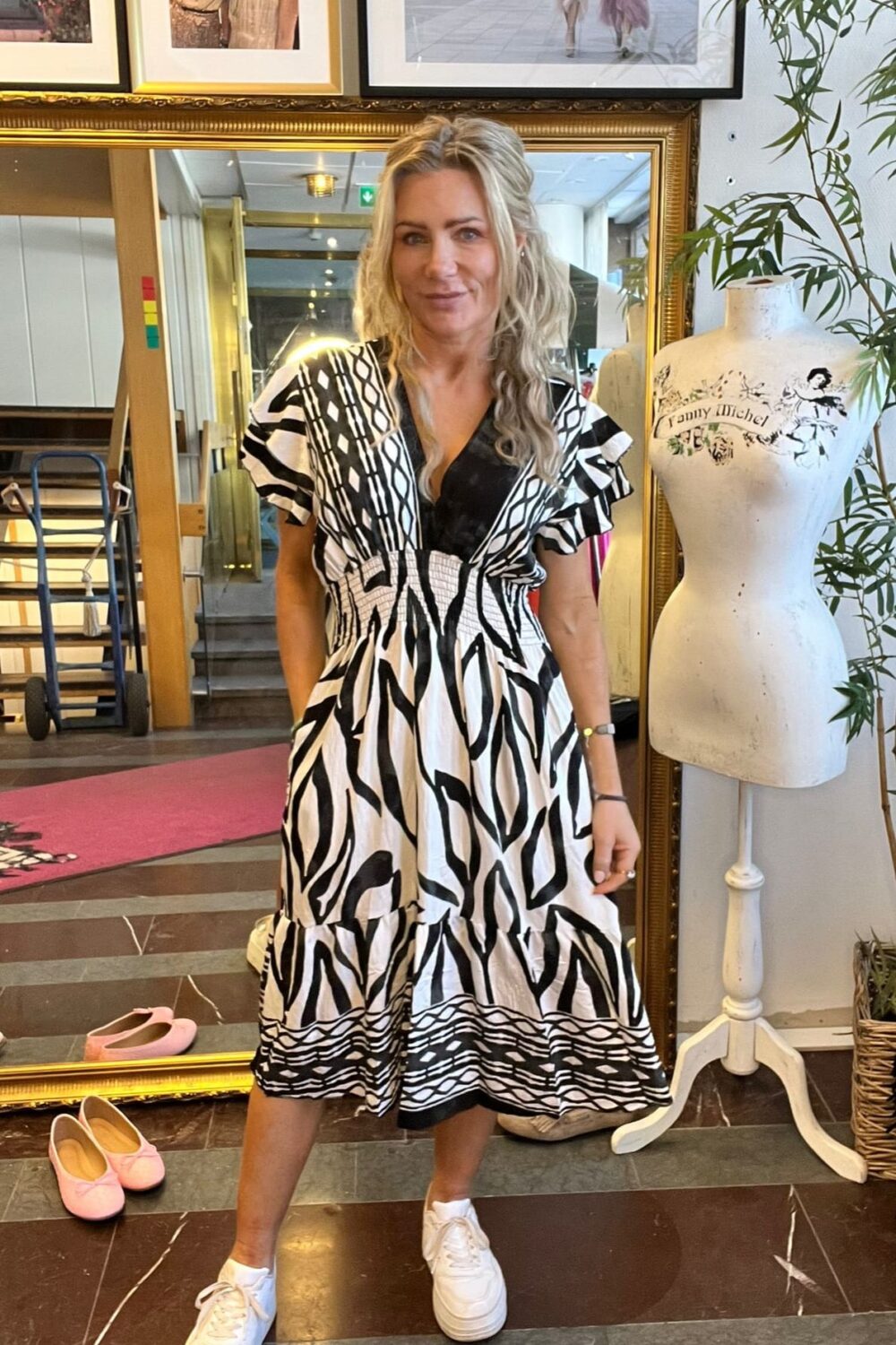 Blond kvinna står och poserar framför en stor guldramsspegel i en butik eller ateljé, iklädd svartvit mönstrad klänning och vita sneakers. Bredvid finns en provdocka med texten “Tommy Michel” och rosa skor på golvet.