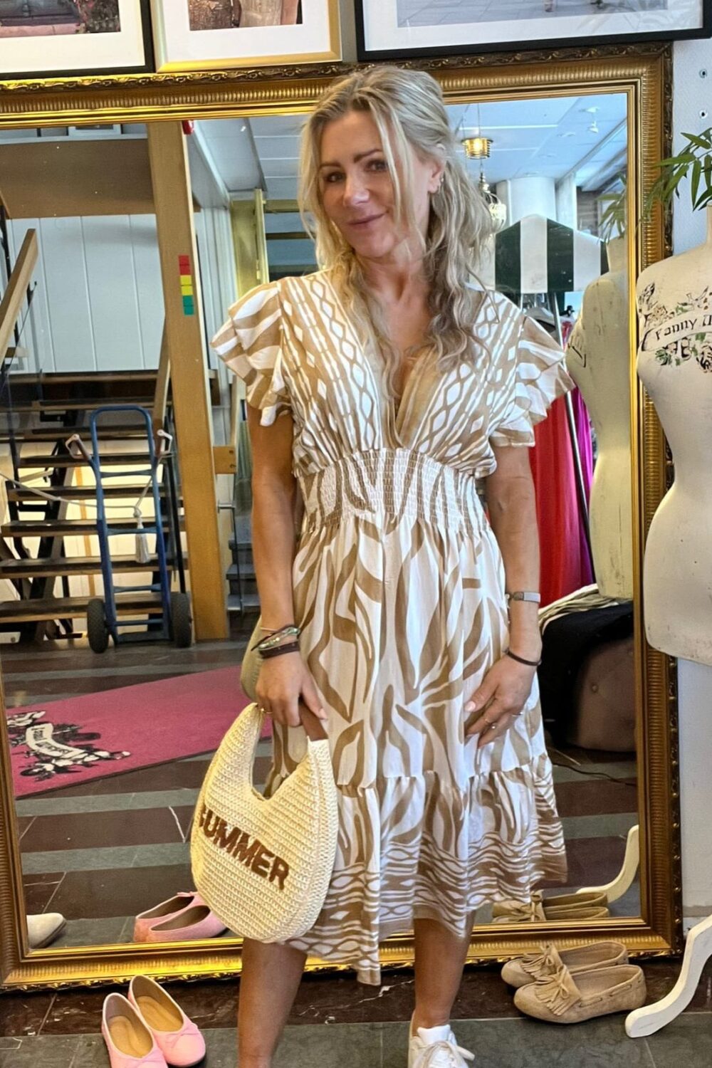 Blond kvinna poserar framför en stor guldramsspegel i en butik, iklädd beigevit mönstrad klänning och vita sneakers. Hon håller en flätad väska med texten ”SUMMER”; skor och provdocka syns runtom.