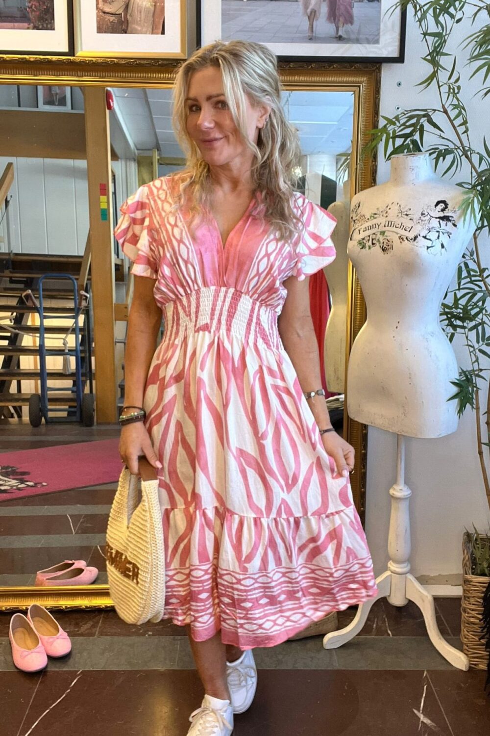 Kvinna i rosa och vit mönstrad klänning poserar och håller en stråväska i en butik eller studio. Bakom henne syns en stor guldramsspegel, inramade foton och en provdocka med texten “Fanny Michel”.