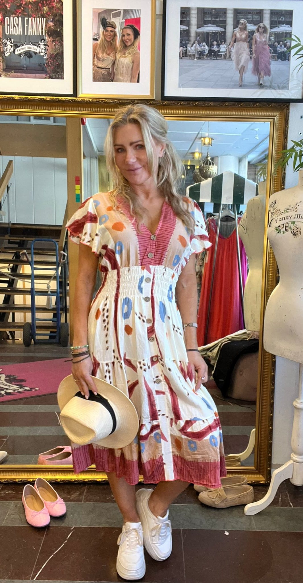 Blond kvinna poserar framför en stor guldramsspegel i en klädbutik, håller en stråhatt och bär mönstrad klänning och vita sneakers. På väggen syns inramade foton och texten “CASA FANNY” samt “Fanny Michel”.