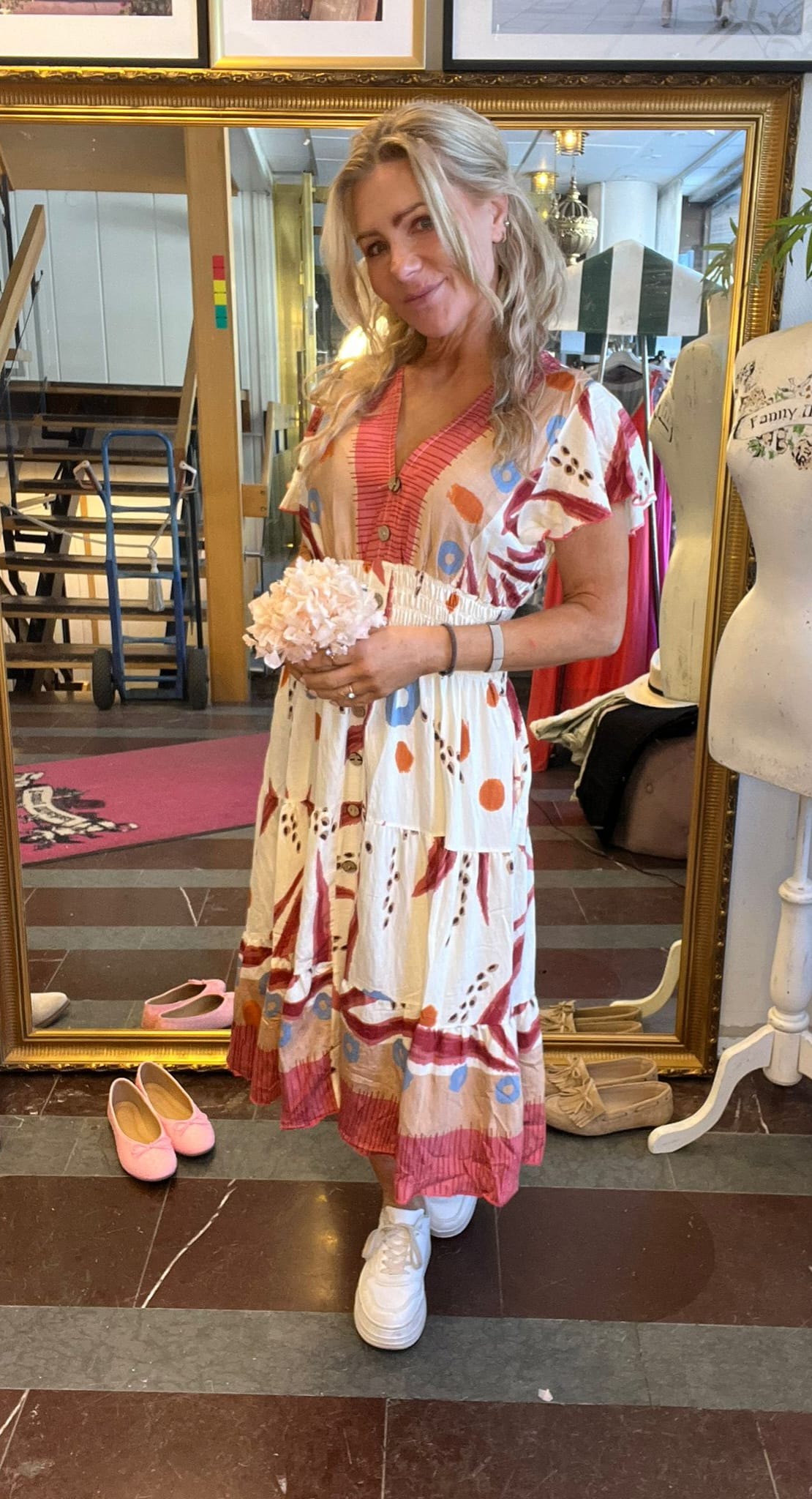 Kvinna med långt blont hår poserar och håller en ljusrosa blomsterbukett framför en stor guldramsspegel. Hon bär en vit mönstrad klänning och vita sneakers i en butik med provdocka och skor.