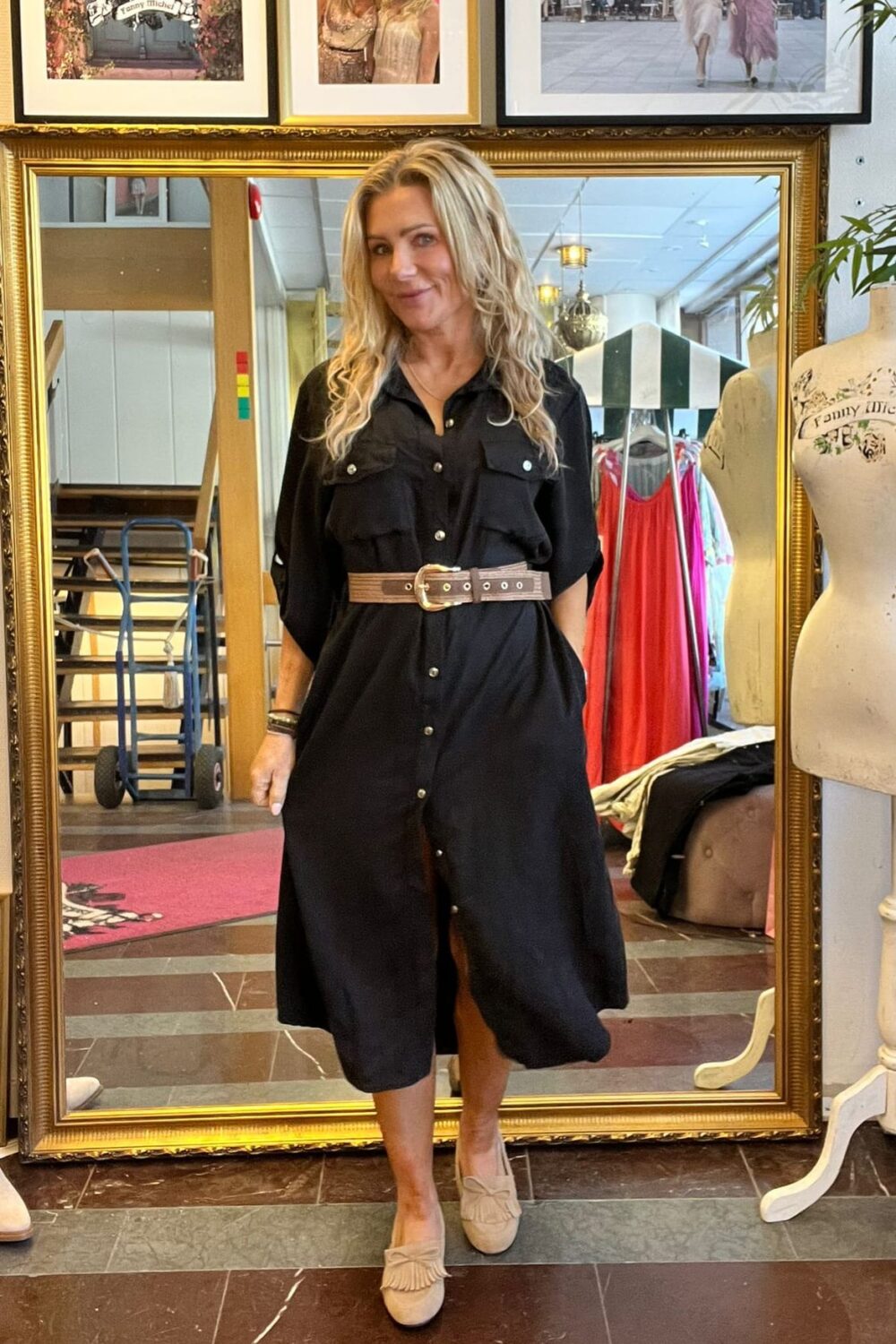 Kvinna med långt blont hår poserar framför en stor guldramsspegel i en klädbutik, klädd i svart skjortklänning med bredt bälte och ljusa skor. I bakgrunden syns klädställning, provdocka och inramade foton.