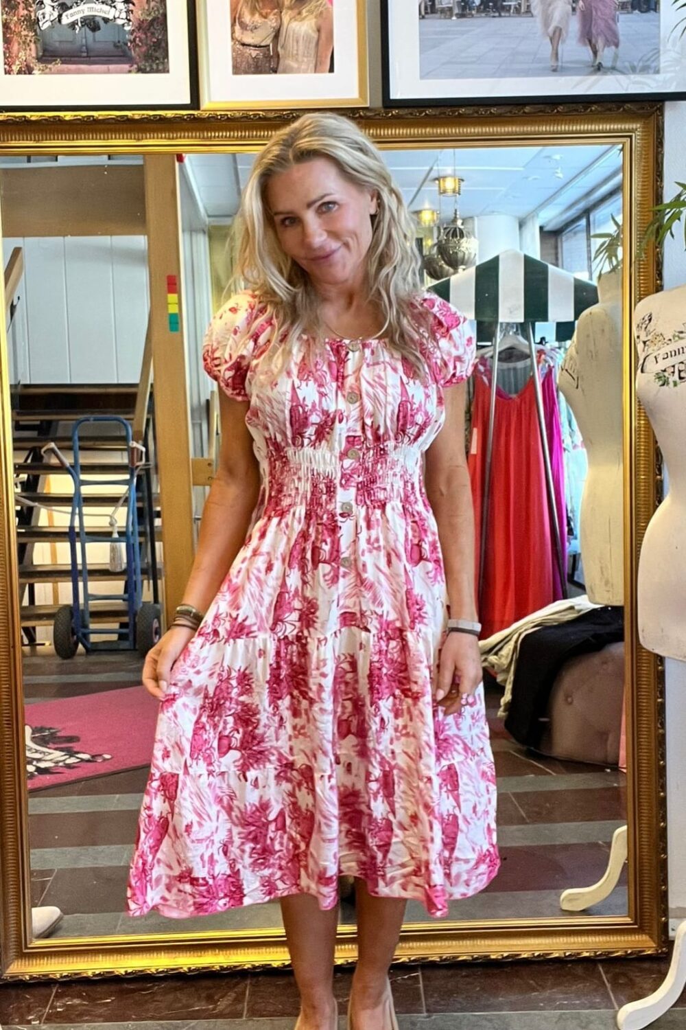 Leende kvinna står framför en stor guldramsspegel och håller ut kjolen på en vit klänning med rosa blommönster. Bakom syns klädhängare, provdocka och inramade foton; ett har texten ”CASA FANNY”.
