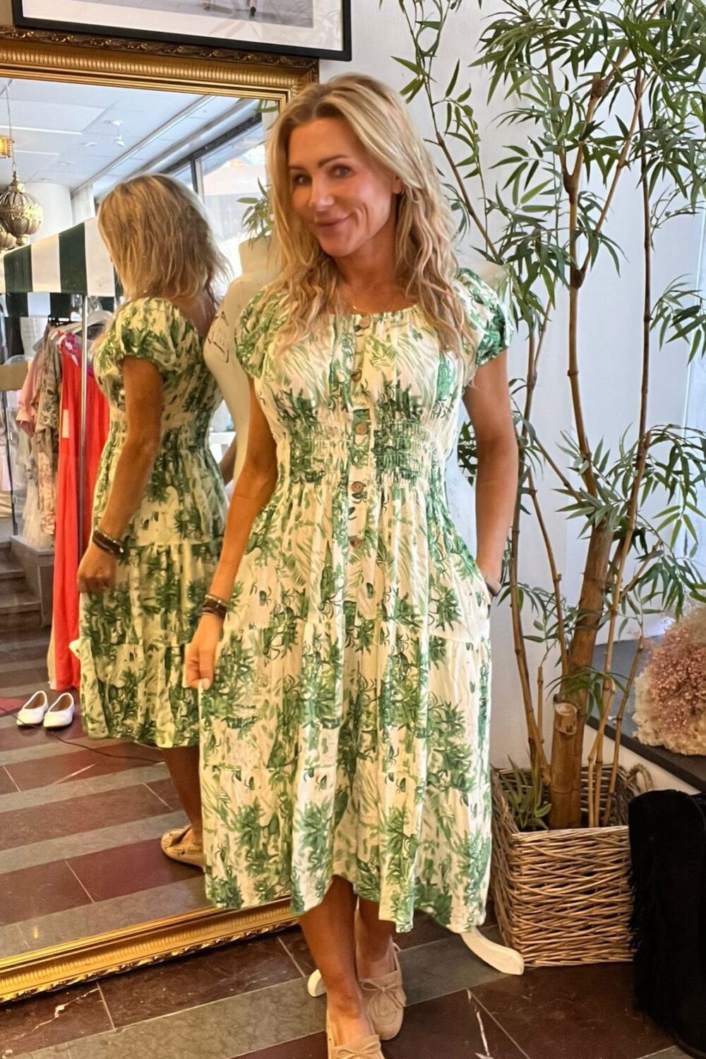 Kvinna står och ler framför en stor spegel, med handen i fickan, och visar en vit klänning med grönt bladmönster och knappar. Hon är i en butik med klädställning, inramad tavla och hög krukväxt.