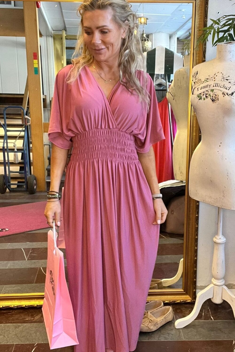 Kvinna i lång rosa klänning står och tittar ner med ett leende, håller en rosa shoppingpåse. Hon är inomhus framför en stor guldramsspegel; bredvid syns en provdocka med texten ”Fanny Michel”.