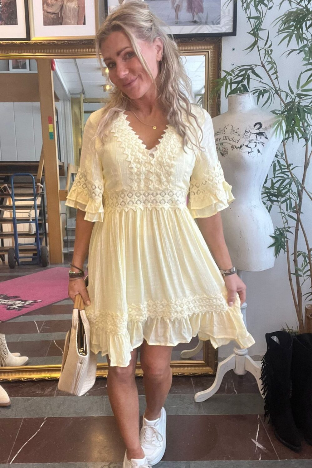 Blond kvinna poserar och ler i en ljusgul, broderad klänning med volanger, håller en vit handväska och bär vita sneakers. Hon står i en butik/studio med spegel, inramade foton, skyltdocka och växt. Text på skyltdocka: “Guy Michel”.