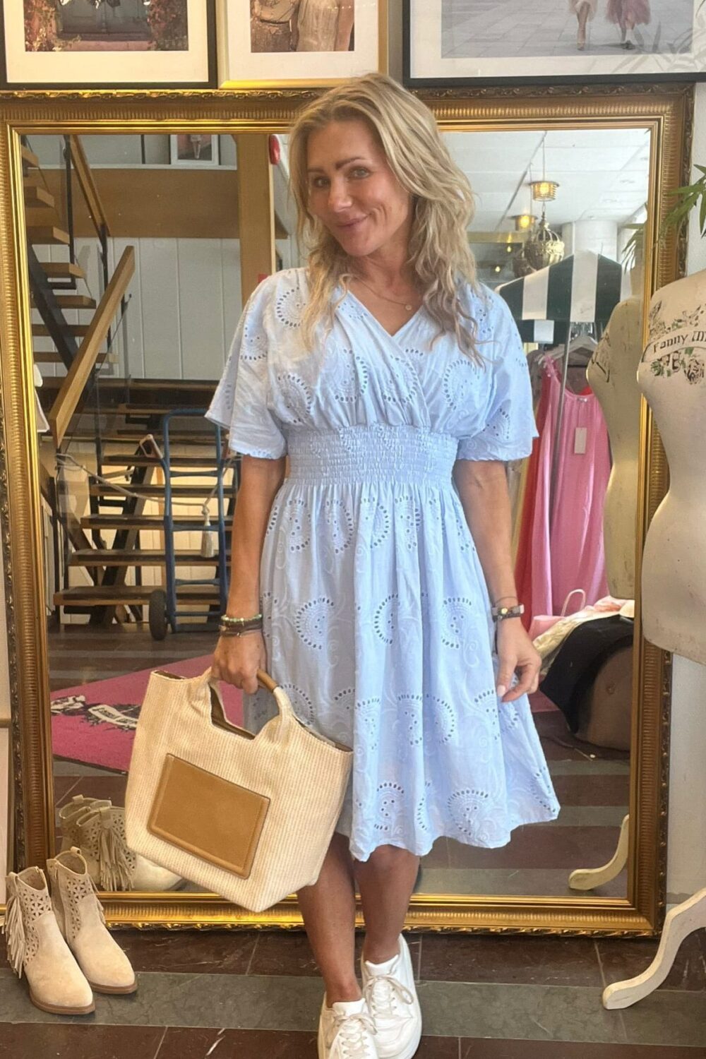 Leende kvinna står framför en stor guldramsspegel och håller en beige tygväska. Hon bär ljusblå broderad klänning och vita sneakers i en klädbutik, med skor på golvet och inramade foton ovanför. Text: “CASA FANNY”.