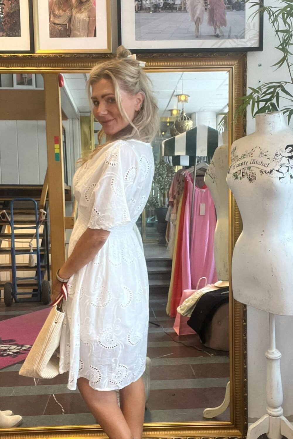 Kvinna i vit broderad sommarklänning står i profil och ler mot kameran i en klädbutik, med vit handväska och sneakers. Bakom henne syns stor guldspegel, hängande klänningar och provdocka med texten “fanny michel”.