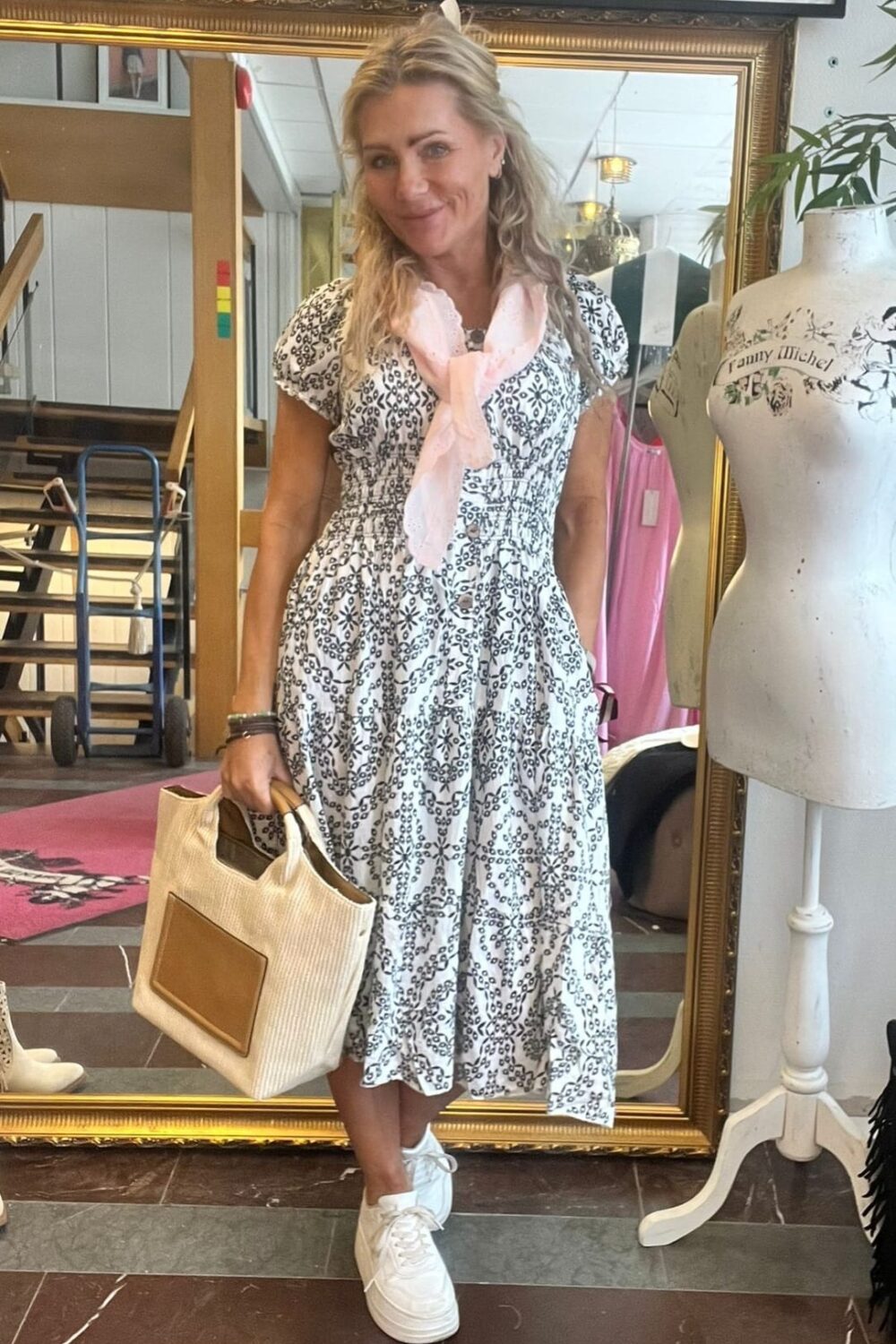 Leende kvinna står framför en stor guldramsspegel och håller en beige handväska. Hon bär vit klänning med svart mönster, ljusrosa scarf och vita sneakers. Bredvid syns en provdocka med texten “Tommy Michel” i butiksmiljö.