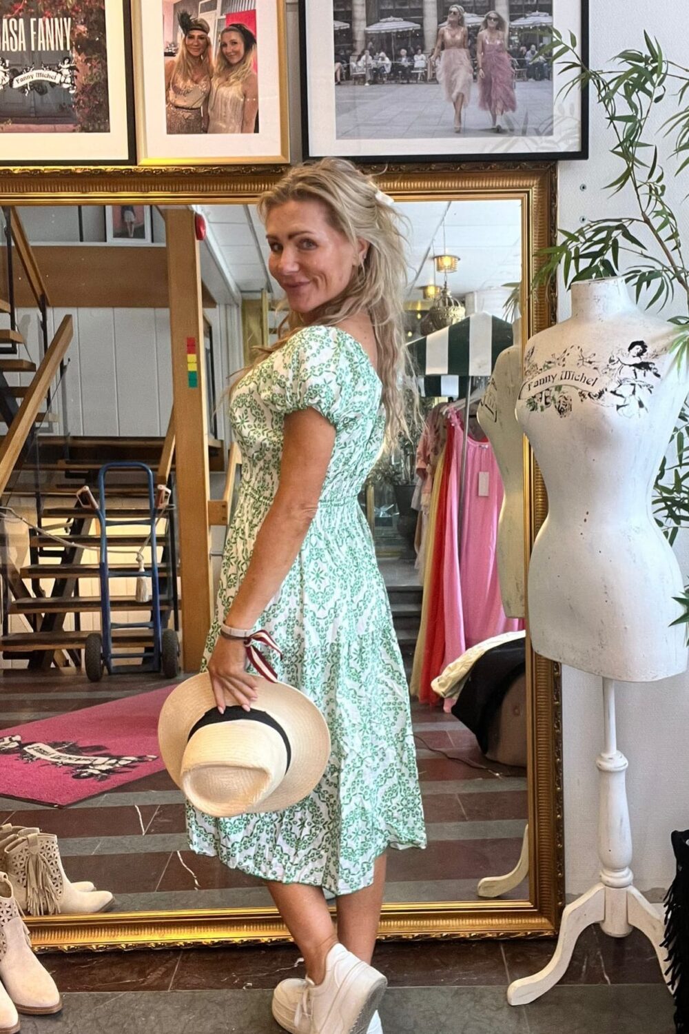 Leende kvinna i grönmönstrad klänning poserar i en butiksmiljö och håller en stråhatt bakom ryggen. Hon står framför en stor guldramsspegel, med klädställning och skyltdocka bredvid samt inramade foton på väggen.