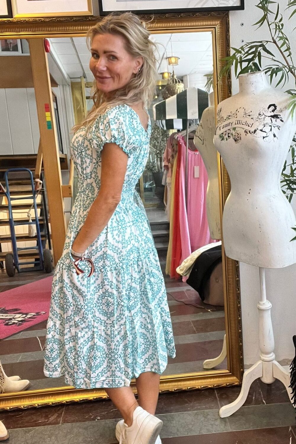 Blond kvinna står i profil och ler mot kameran medan hon visar upp en vit klänning med turkost mönster i en butik. Bakom henne syns en stor guldramsspegel, klädställning, inramade foton och provdocka med texten ”Tommy Hilchel”.