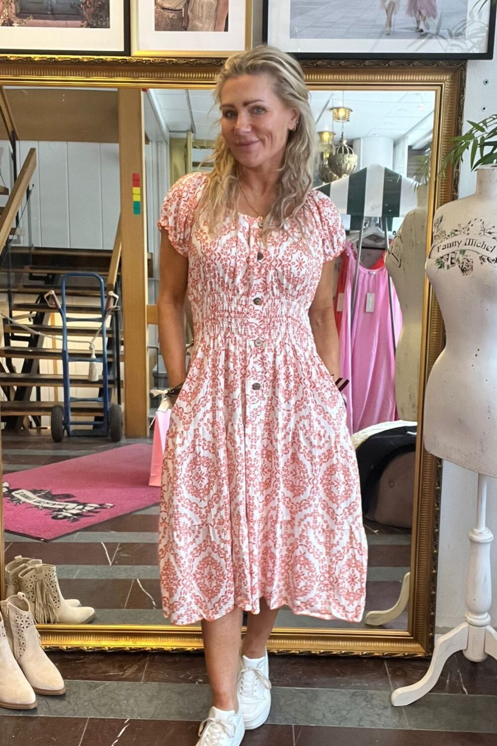 Blond kvinna står och ler med händerna i fickorna, klädd i vit klänning med rött mönster och vita sneakers, framför en stor spegel i en butik. På väggen syns inramade foton och texten ”CASA FANNY” och ”Fanny Michel”.