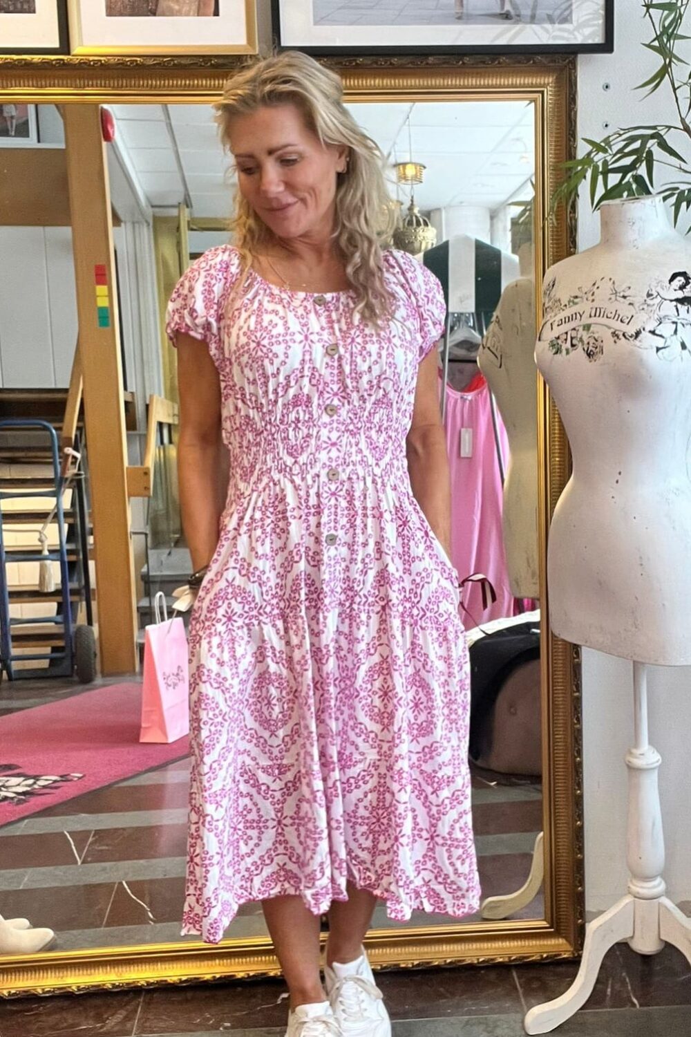 Blond kvinna står framför en stor spegel och tittar ned med ett leende, klädd i vit klänning med rosa mönster och vita sneakers. Bredvid syns provdocka med texten “Fanny Michel” och inramade foton på väggen.