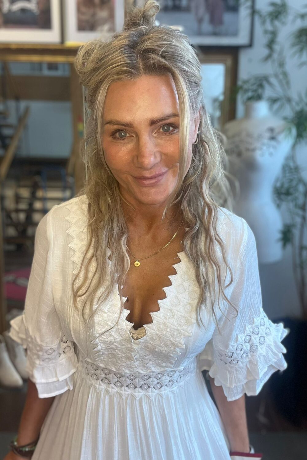Leende kvinna med långt blont hår tittar in i kameran och poserar inomhus. Hon bär en vit broderad klänning med volangärmar och ett halsband med bokstaven A, med tavlor och grön växt i bakgrunden.